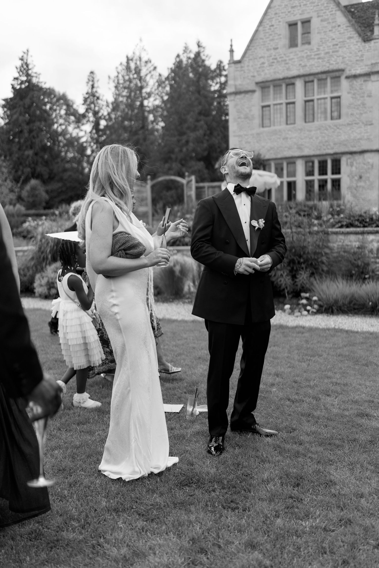 Bride in white gown and groom in tuxedo laugh together on lawn outside stone manor wedding venue.