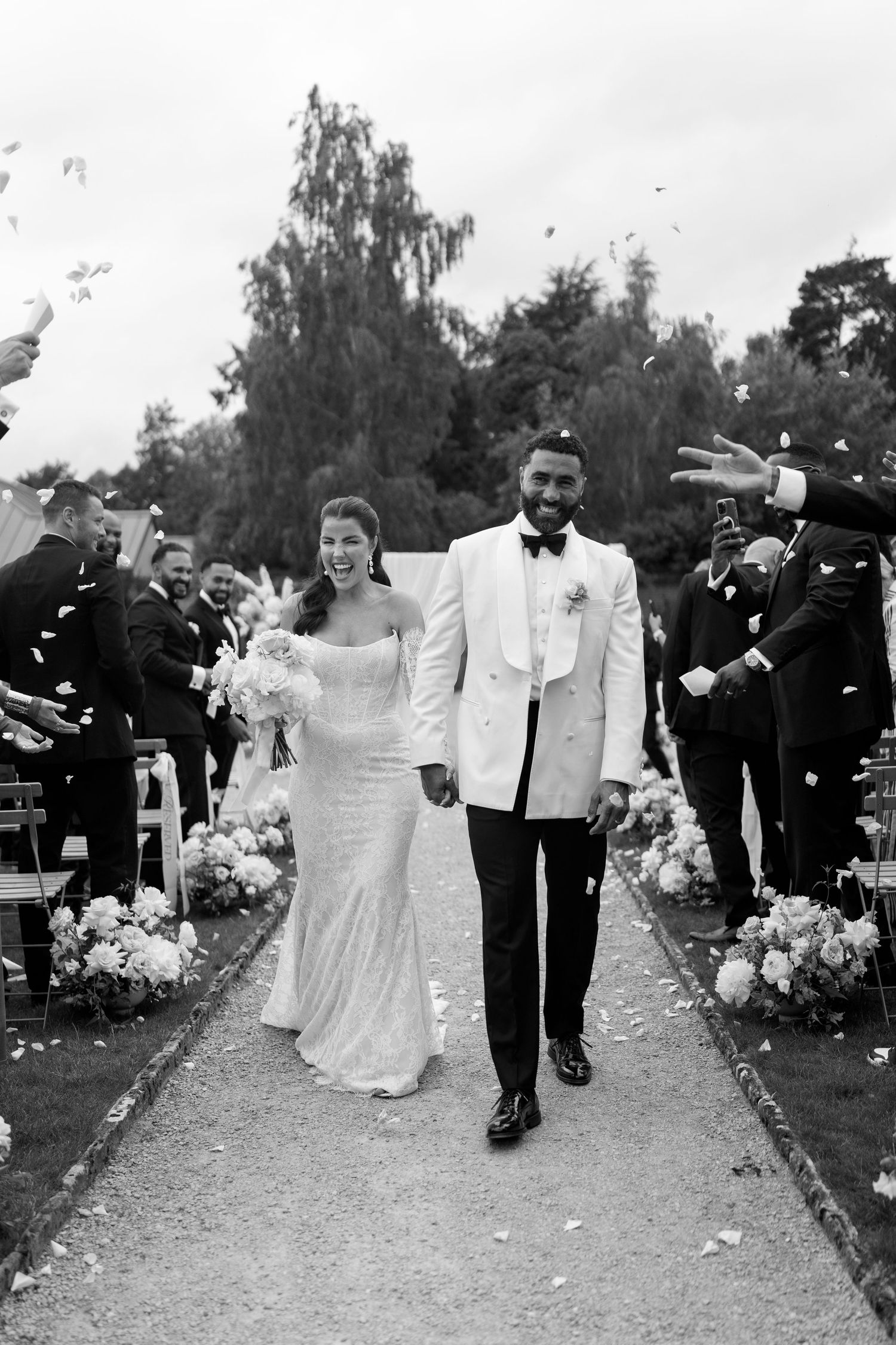 Newlyweds walk down aisle as guests throw confetti at outdoor garden wedding ceremony in black white.