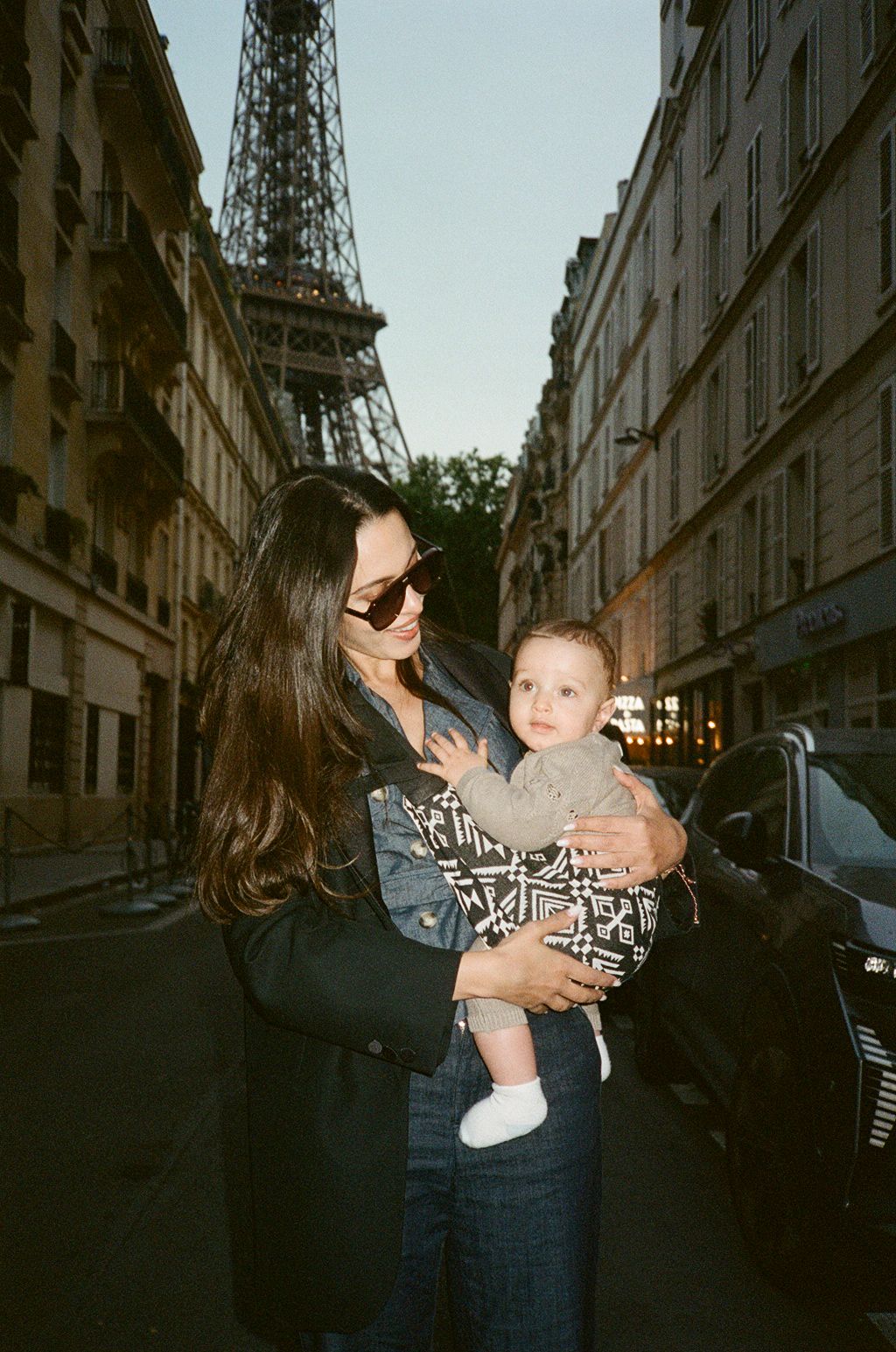 Mother holding baby with Eiffel Tower backdrop on Parisian street.