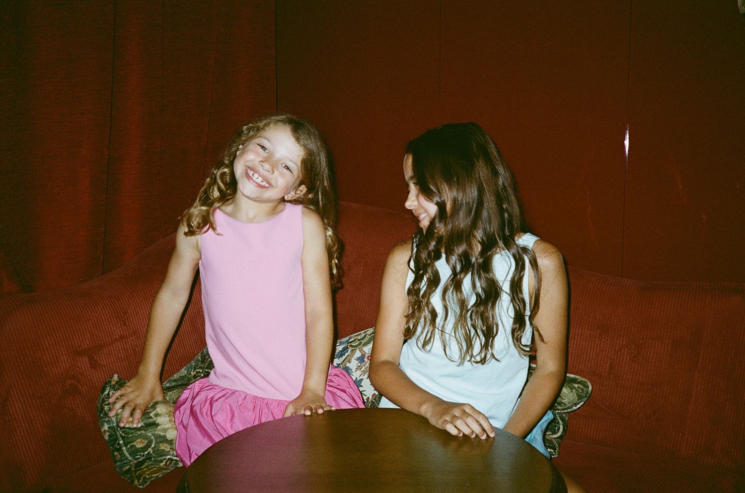 Two young girls smiling and chatting on a red sofa, one in pink outfit and one in white top.