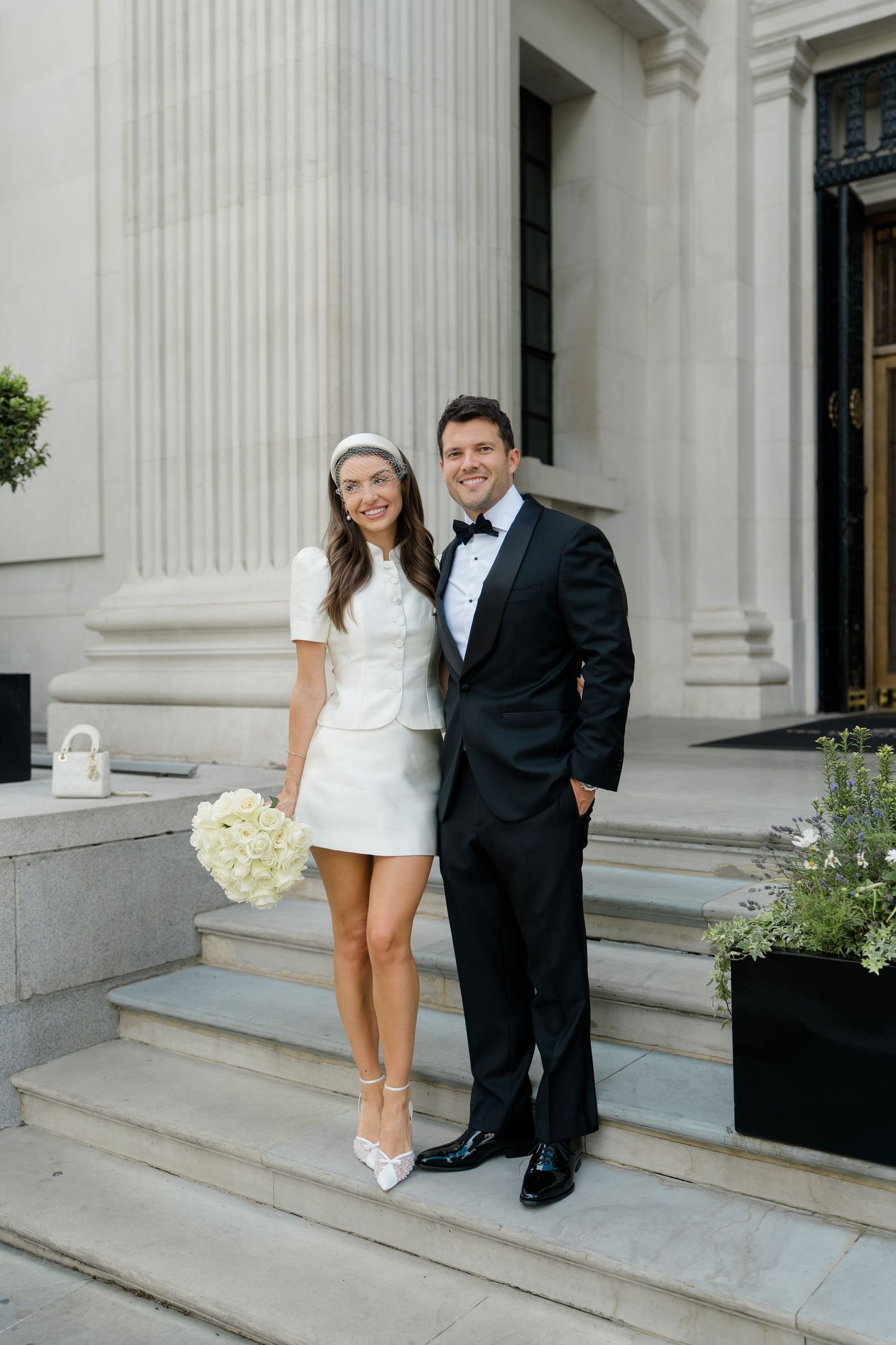 Bride in white mini dress and groom in black tuxedo pose on stone steps outside elegant building.