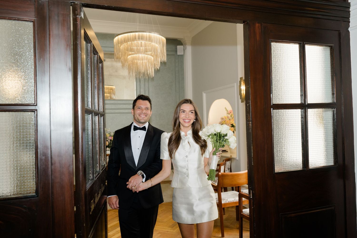 Elegant bride and groom entering a grand room with chandelier.