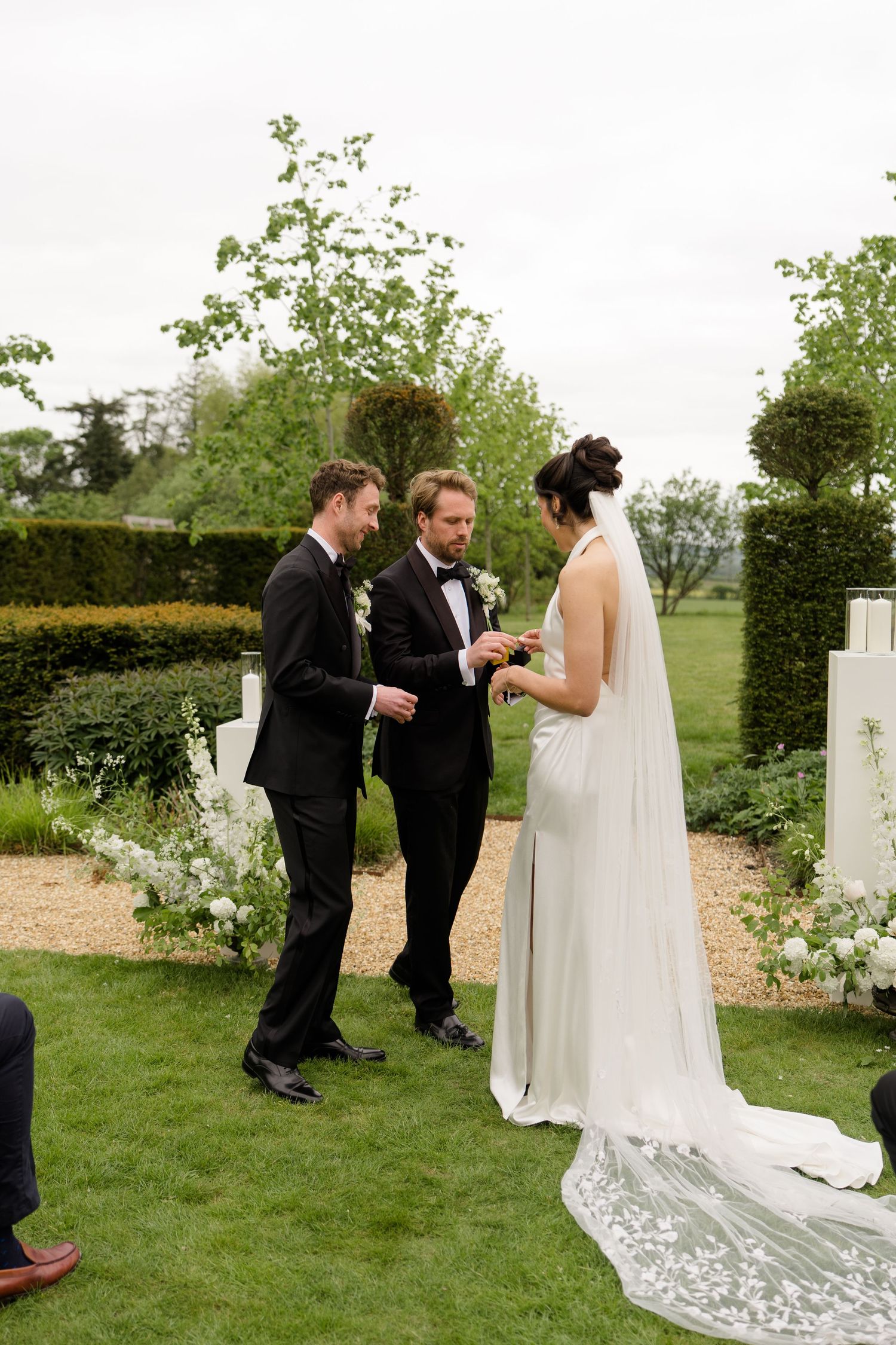 Bride and groom exchange rings during an elegant outdoor wedding ceremony in a lush garden setting.