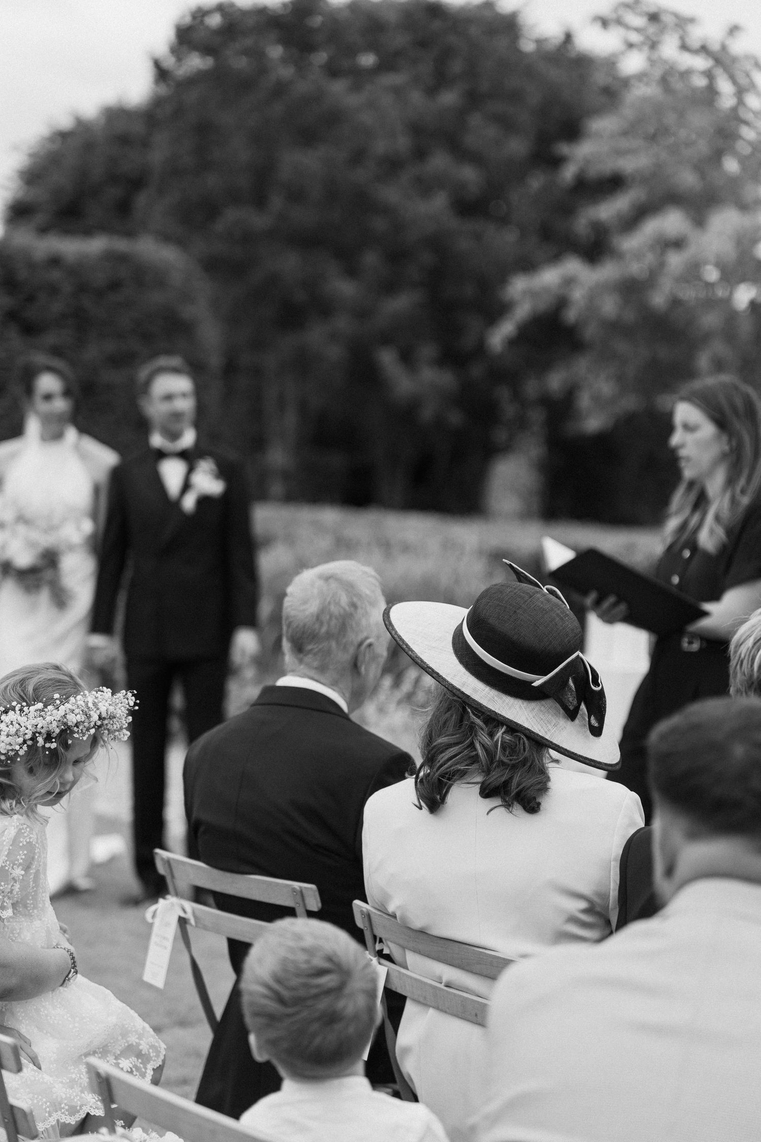 Wedding guests seated outdoors watching ceremony, woman in elegant wide-brim hat in foreground.
