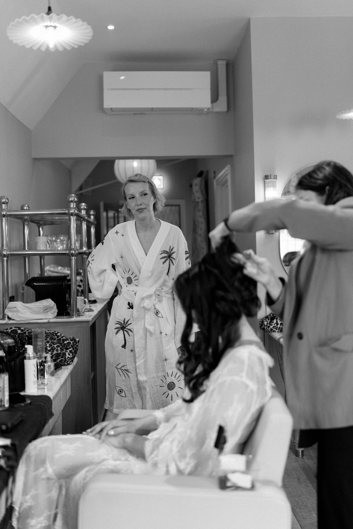 Black and white photo of bride in floral robe watching bridesmaid getting hair styled in mirror reflection.