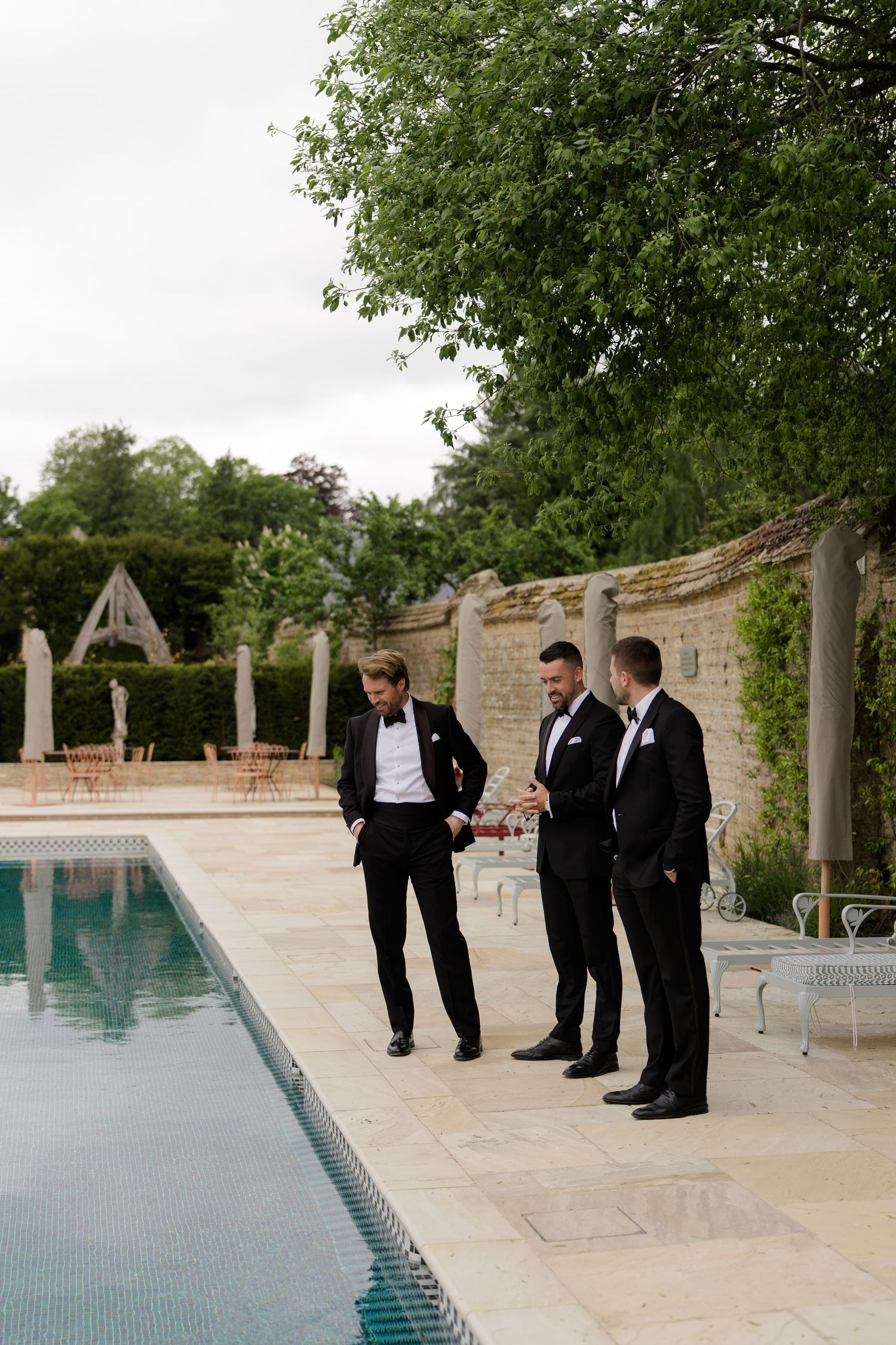 Three groomsmen in black tuxedos standing beside a pool in a manicured garden venue on a cloudy day.