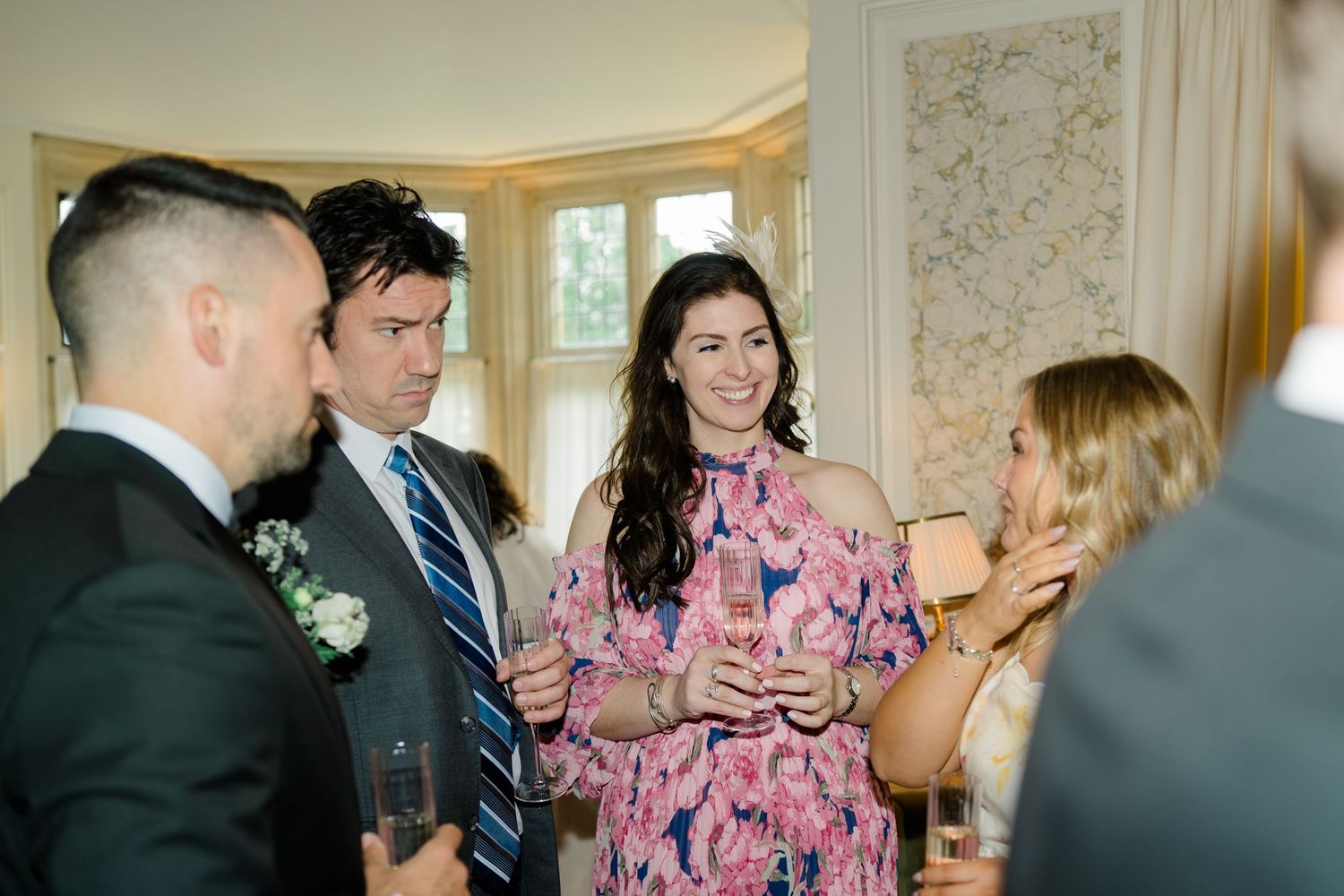 Wedding guests in formal attire socializing indoors, woman in pink floral dress holding champagne.