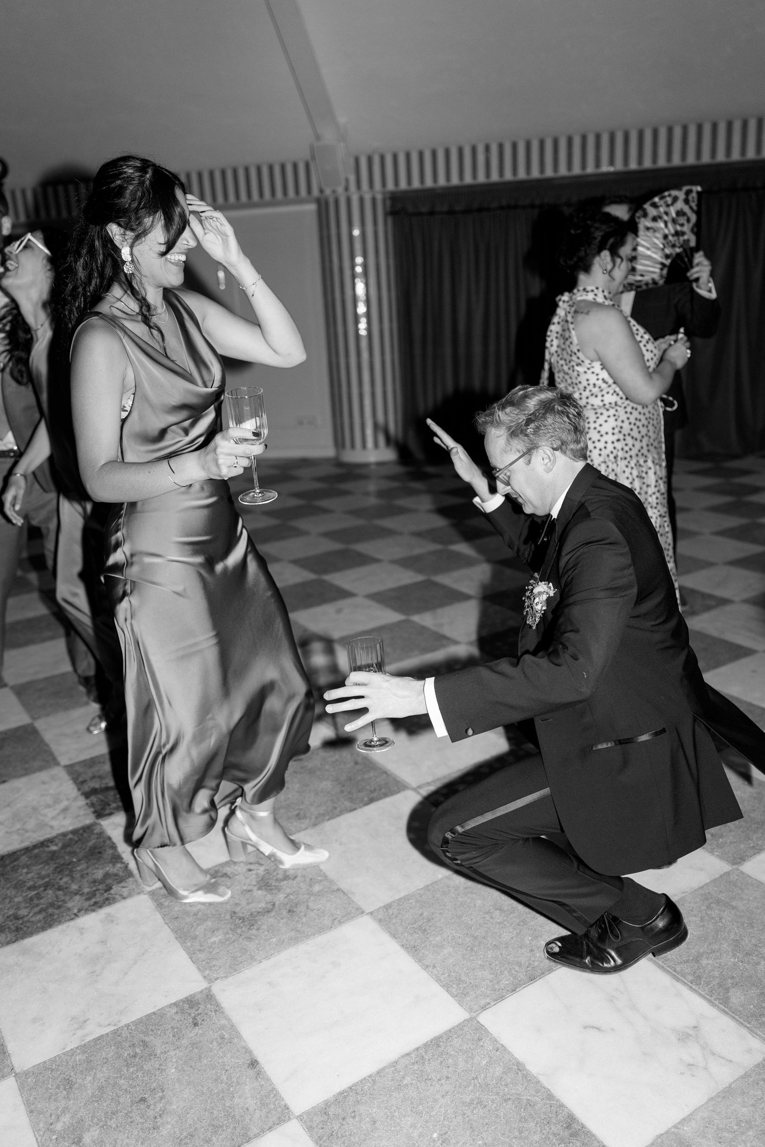 Wedding guest kneels on checkered dance floor in playful dance move during lively wedding reception.
