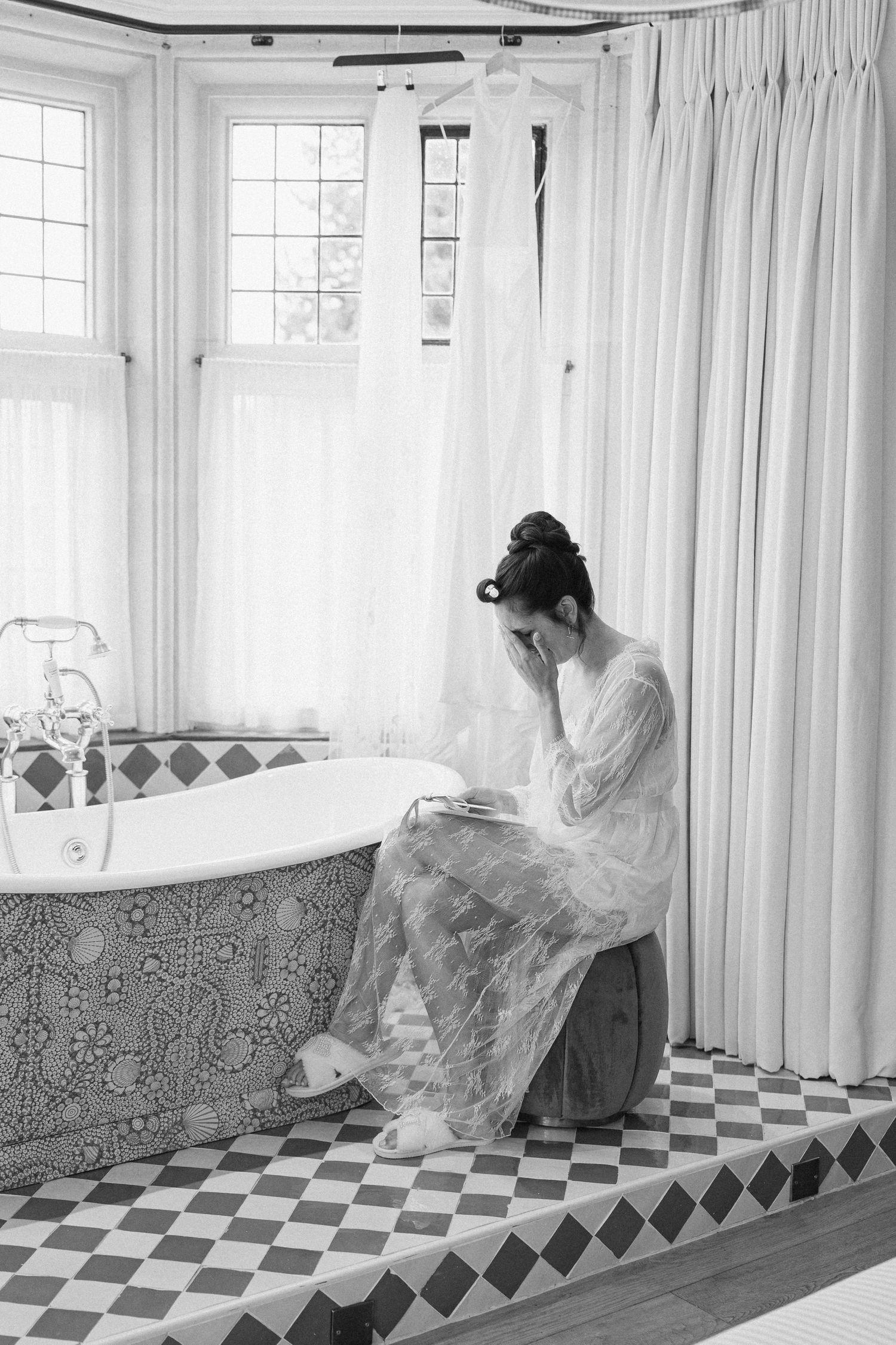 Bride in lace robe sits pensively by ornate clawfoot tub on checkered tile floor in elegant bathroom.