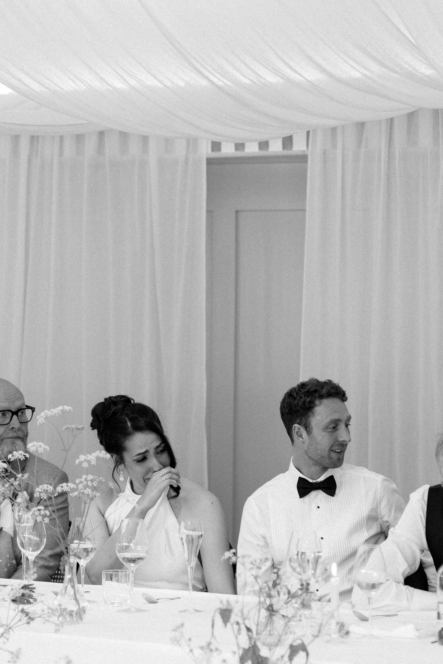 Black and white photo of bride and groom laughing at head table during wedding speeches.