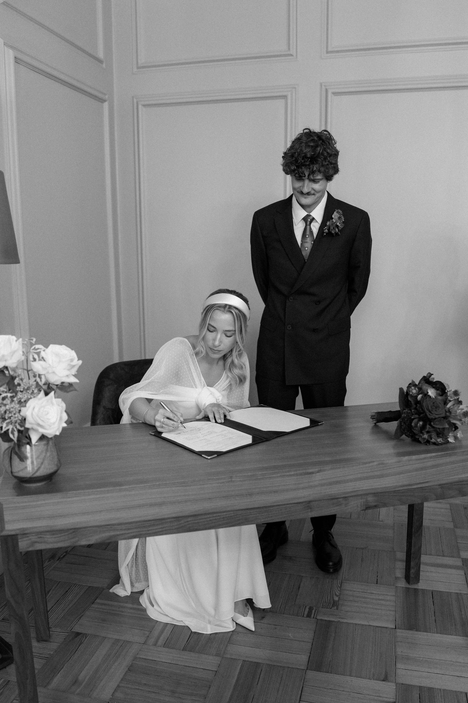 Wedding ceremony participants signing documents at a long wooden table with floral decorations.