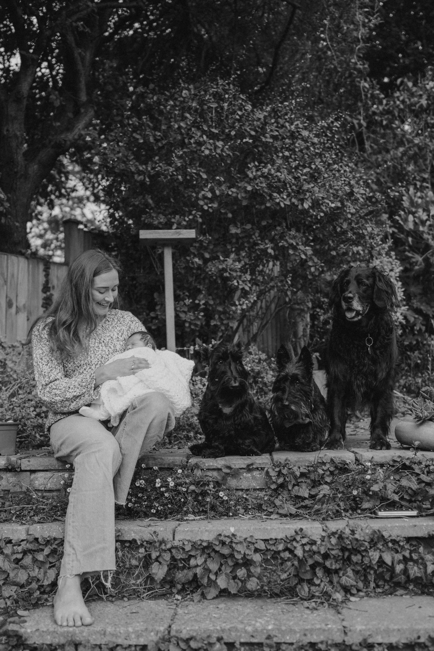 Black and white image of someone sitting on a stone wall in a garden setting.