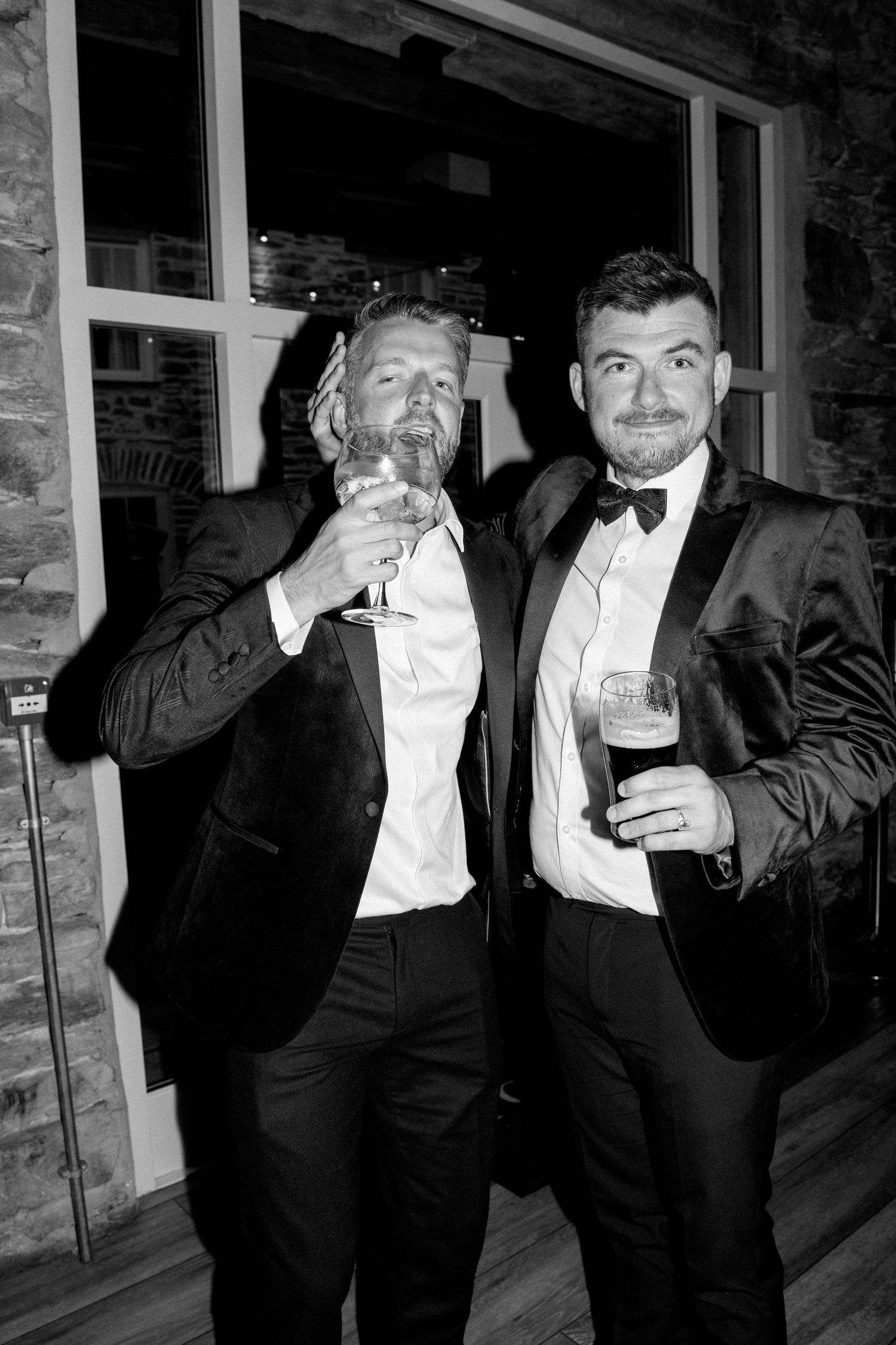 Guests in formal black attire holding drinks and posing against a stone wall background.