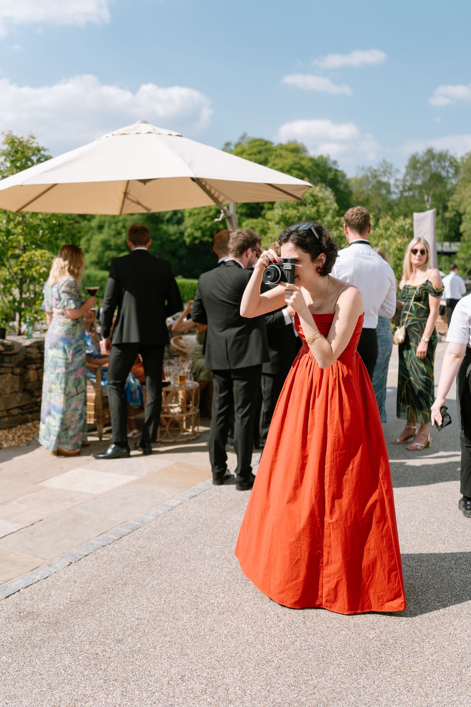 Wedding guests in formal attire gather under beige umbrellas at an outdoor reception.