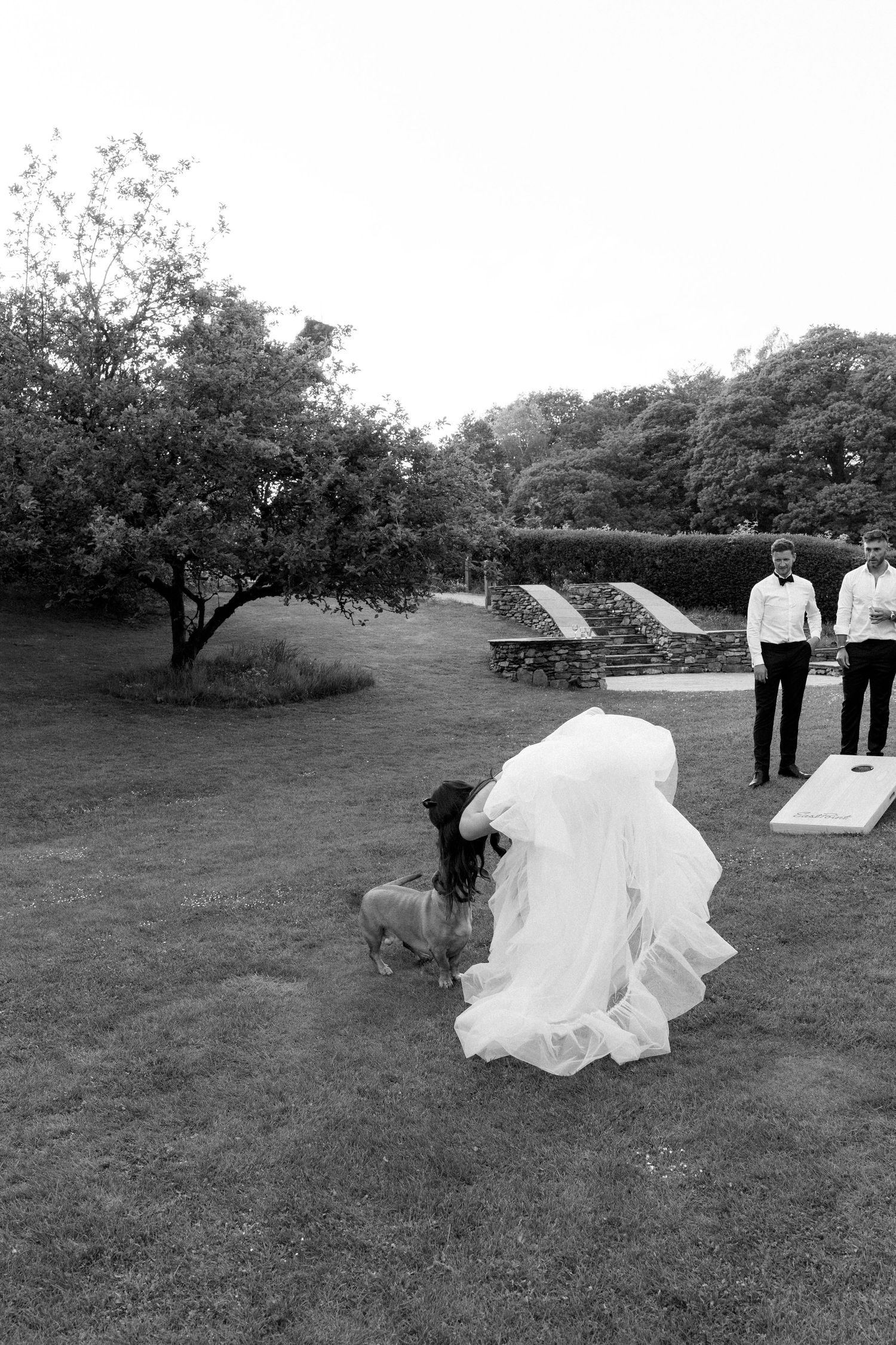 People in formal wedding attire interact playfully on grass with white decorative elements.