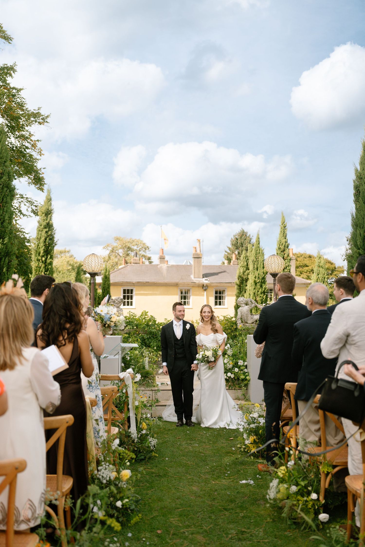 Outdoor wedding ceremony setup with wooden chairs and floral decorations under blue sky.