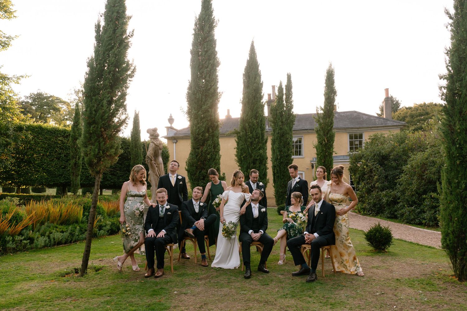Panoramic view of wedding party gathered outdoors with cypress trees and buildings in background.