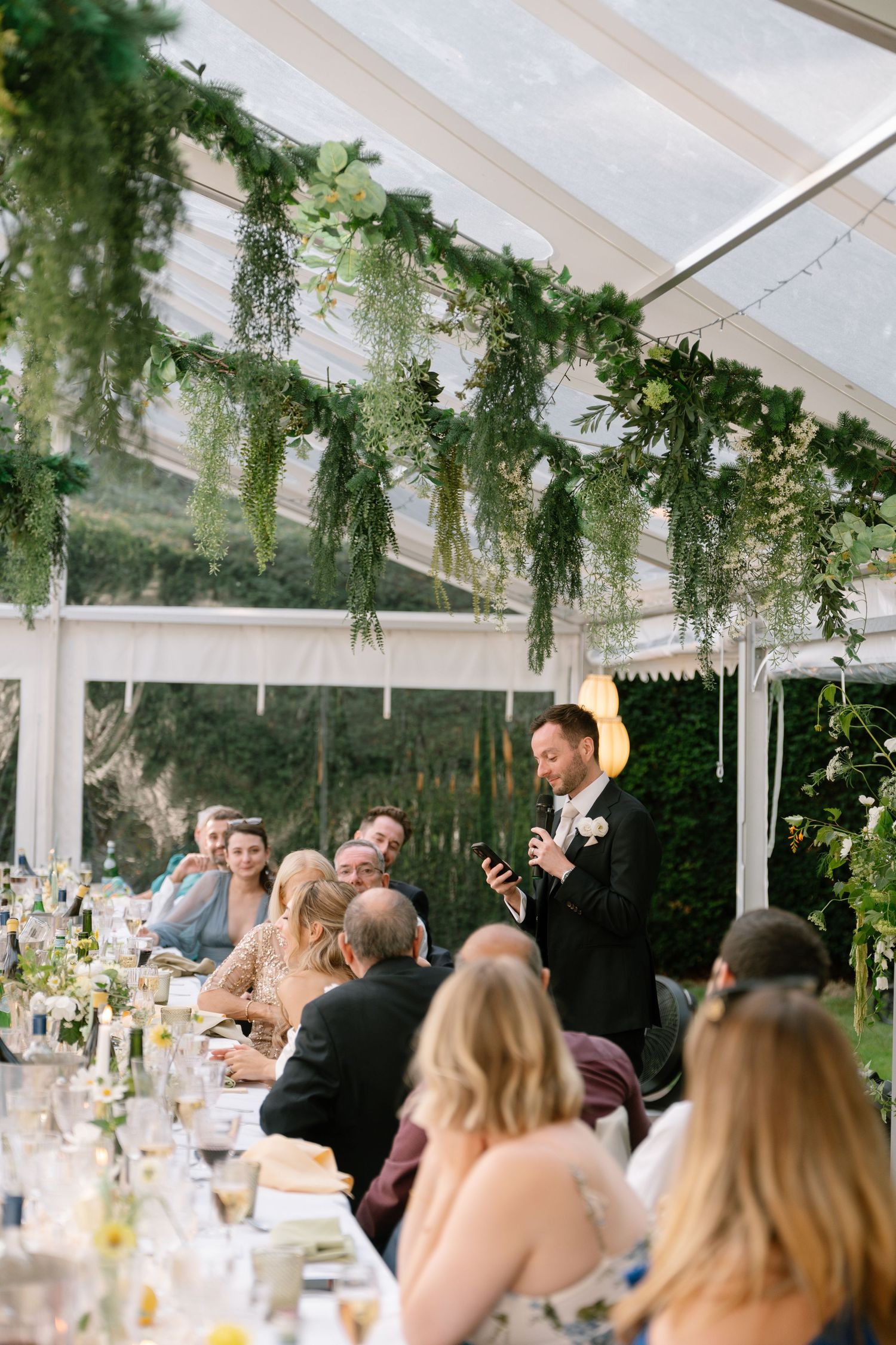 Outdoor wedding reception under tent with hanging greenery and white decor.