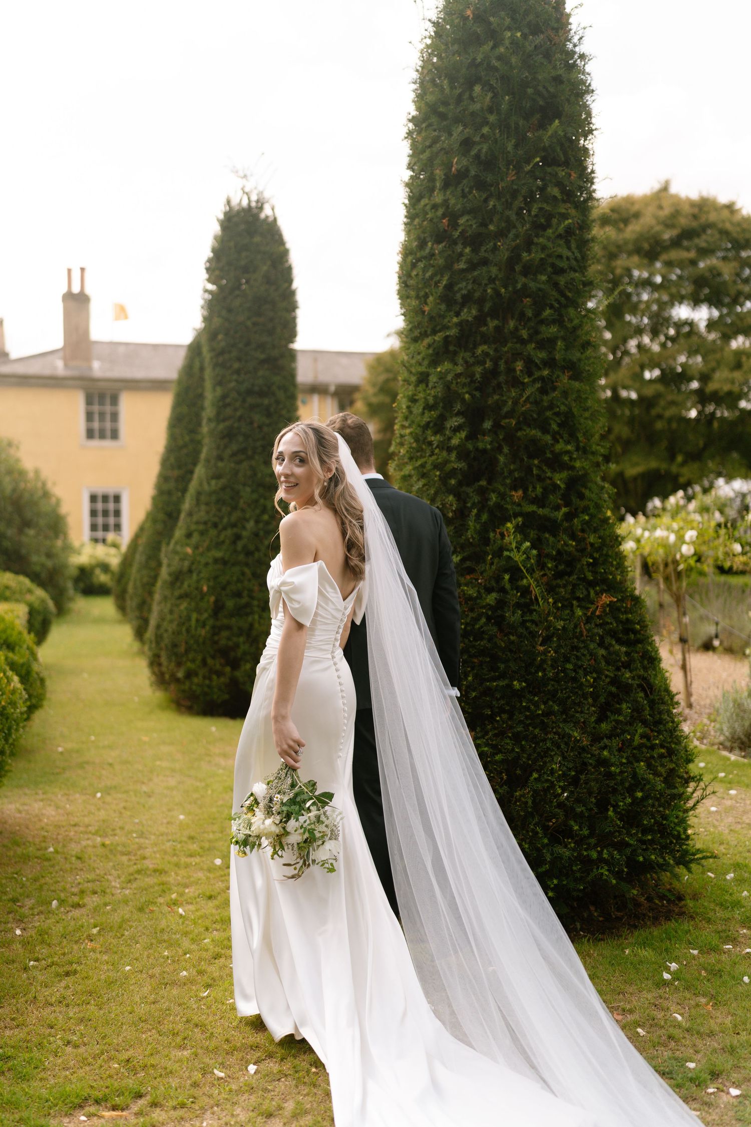 Newlyweds walking through a garden path lined with conical evergreen trees.