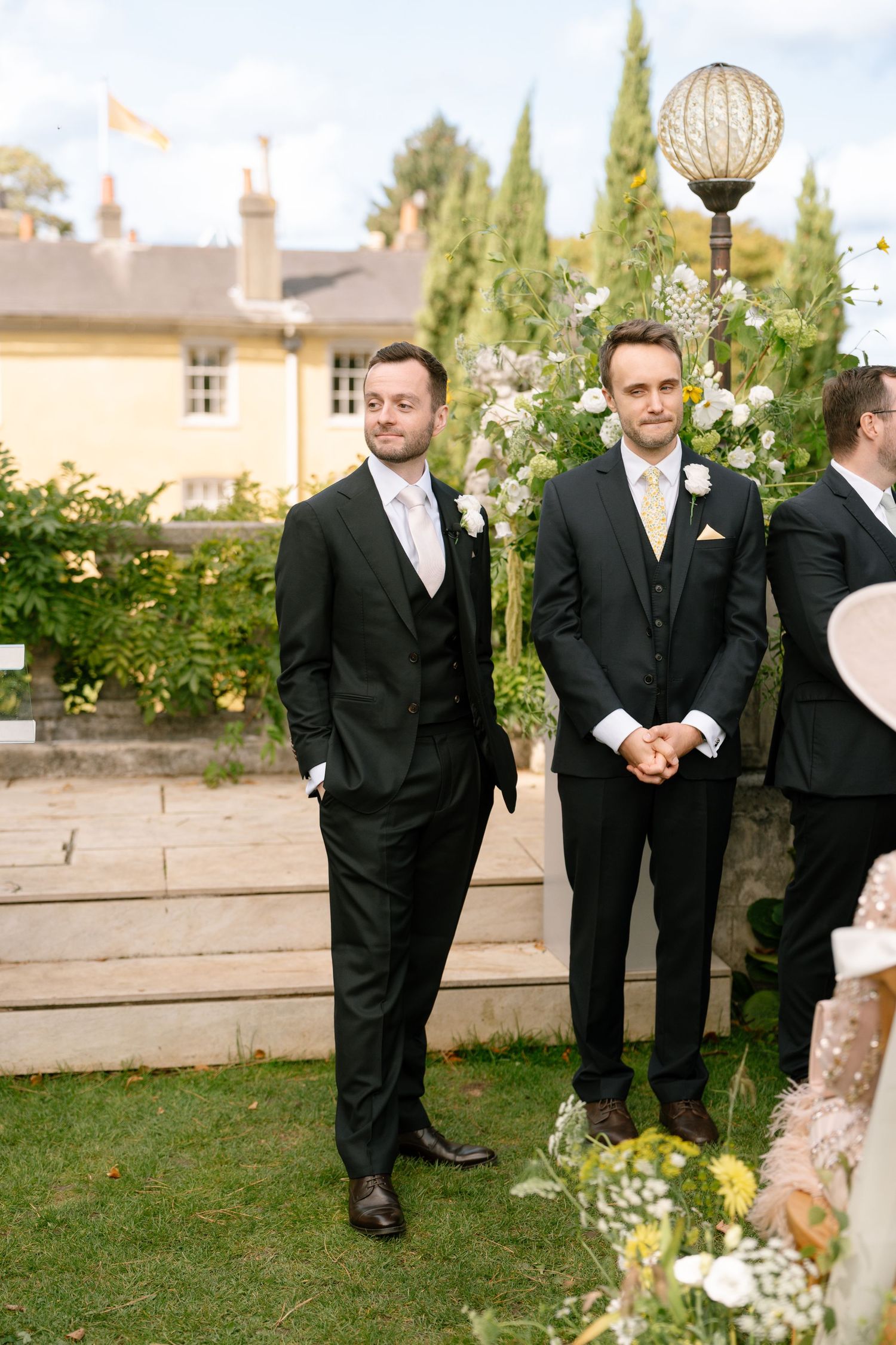 Groomsmen in black tuxedos standing on manicured lawn with garden decor and cypress trees.