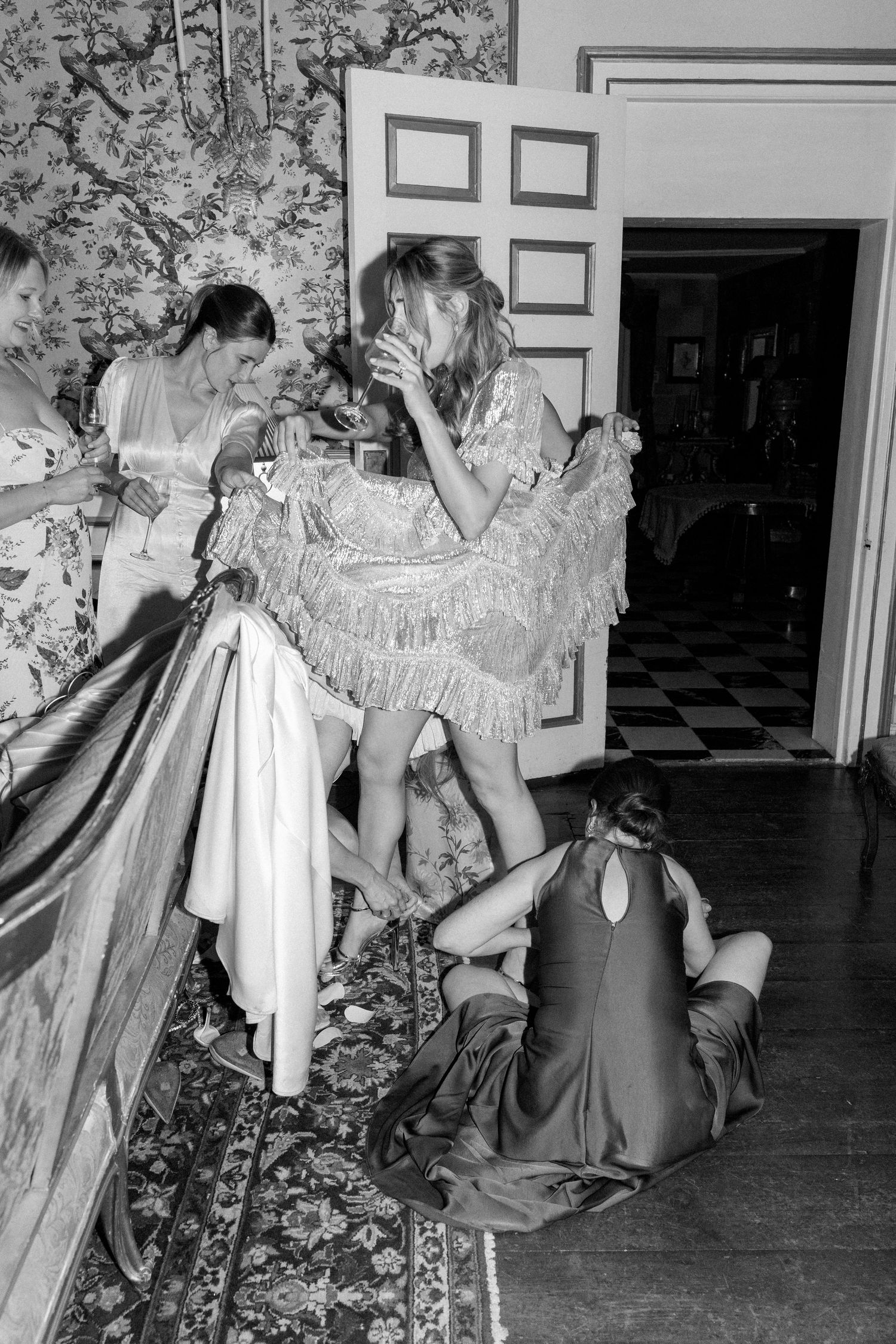 Group gathered in period costumes against floral wallpaper in an elegant interior setting.
