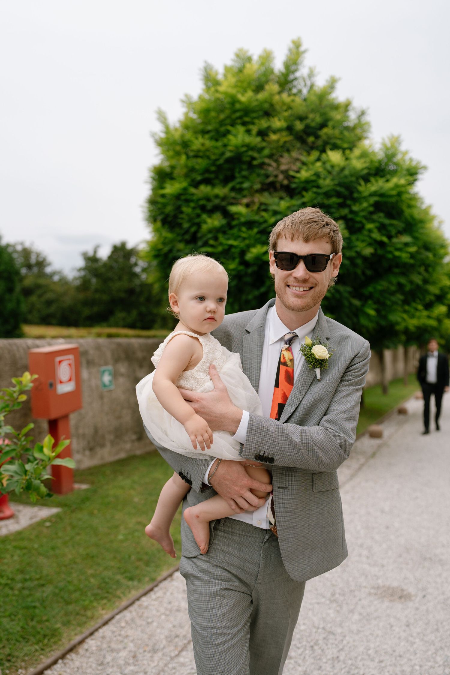 Person in gray suit holds small child in formal wear at outdoor wedding venue.