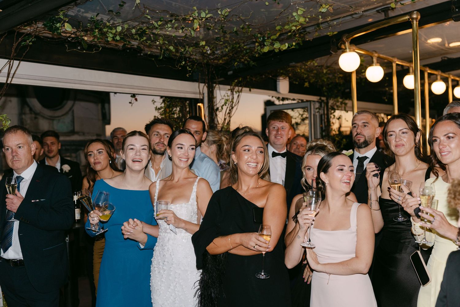 Wedding guests gather under decorative vines and string lights during an evening celebration.