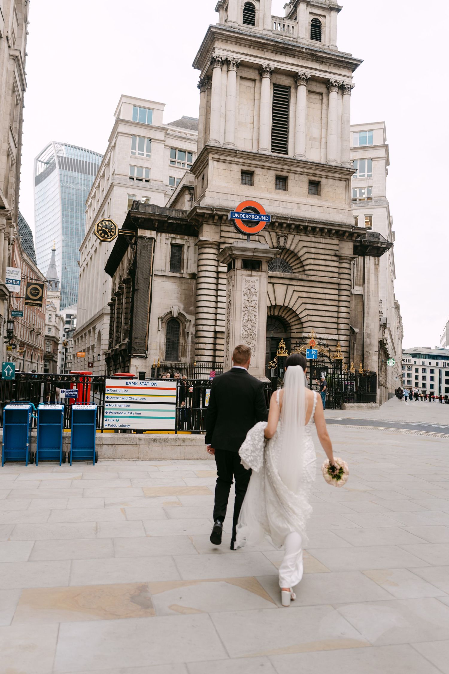 Newlyweds walking towards a London Underground station entrance in formal wedding attire.