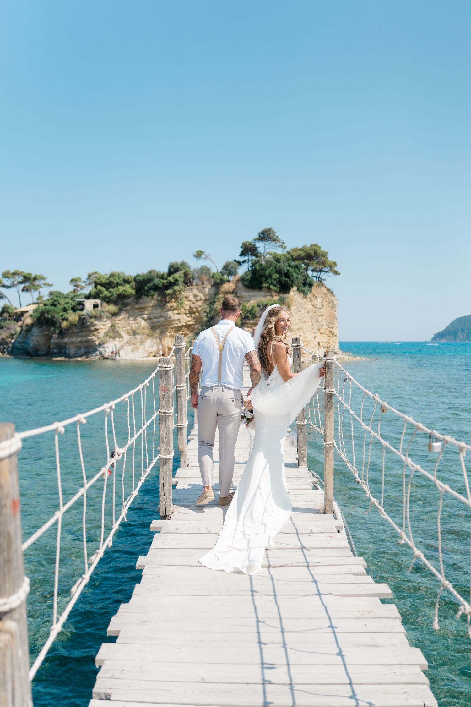 People in white attire walking on a wooden pier over crystal blue Mediterranean waters.