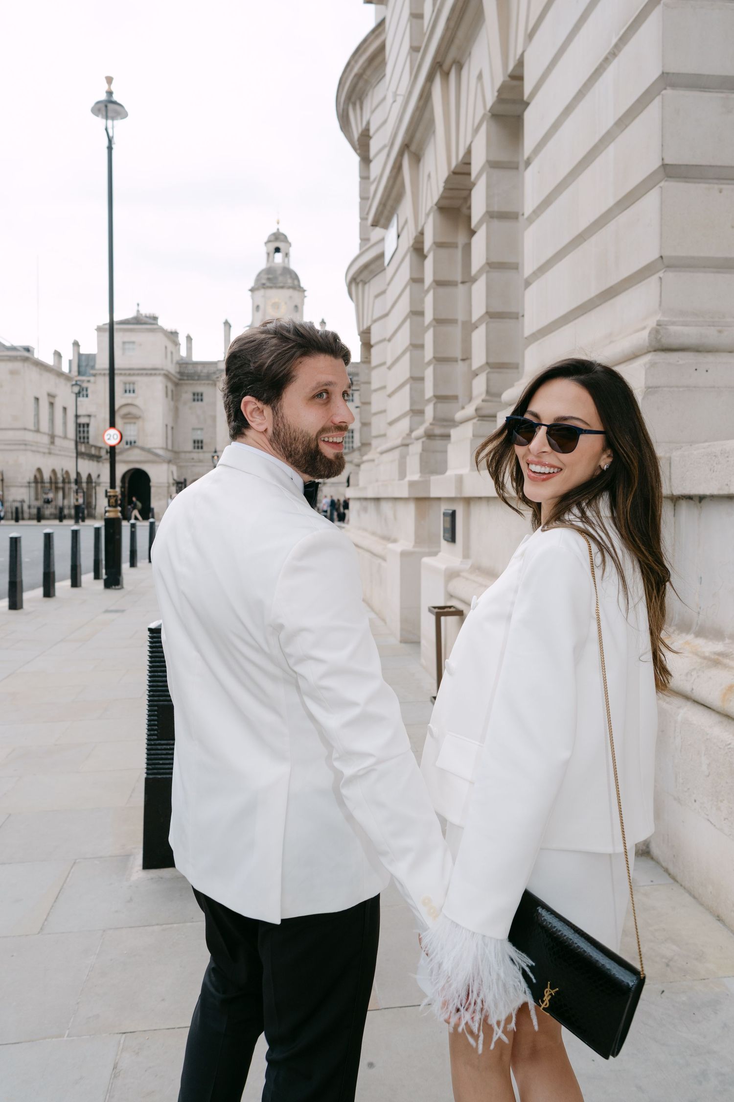 A stylish couple in white outfits poses together on an elegant city street.