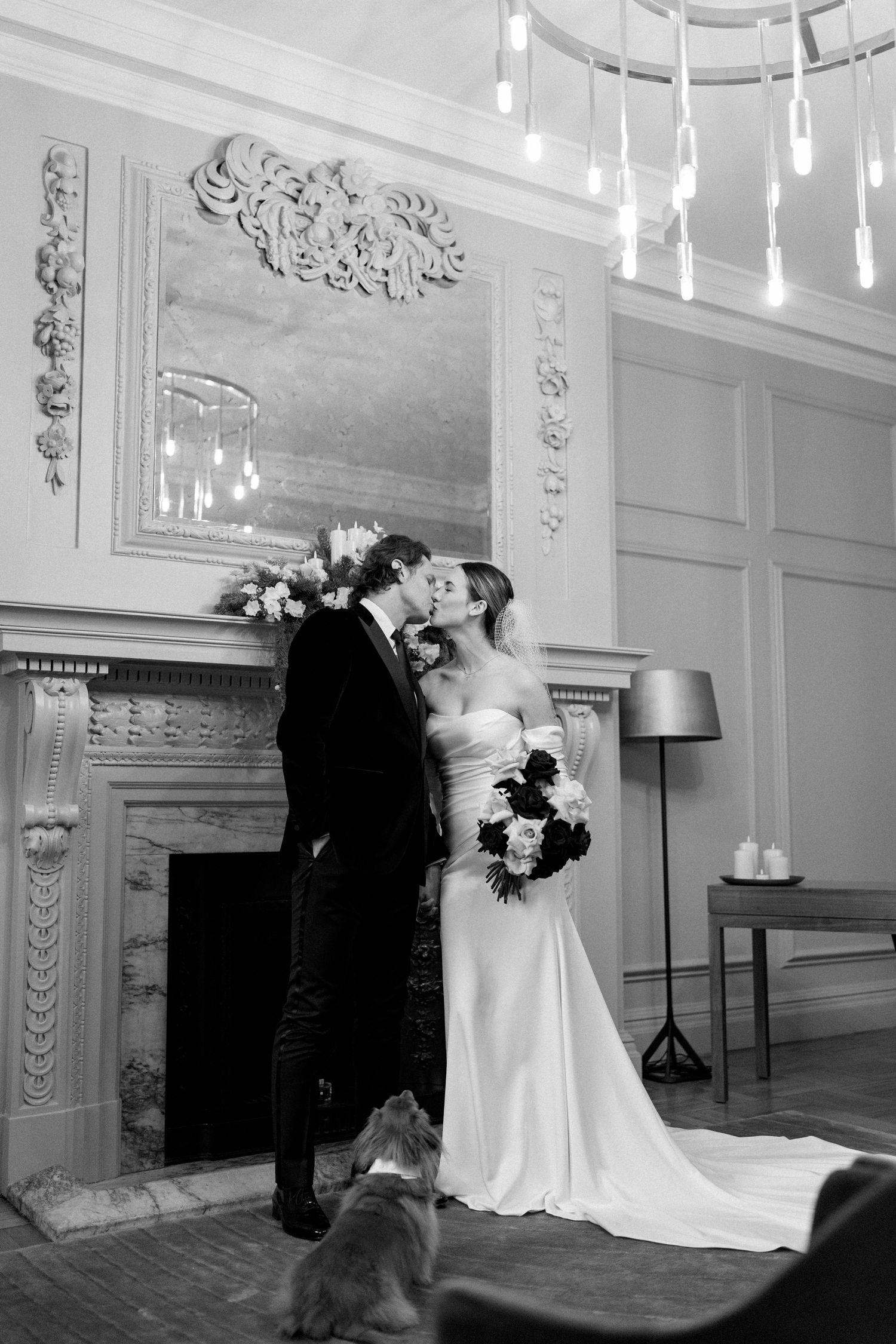 A black and white wedding portrait by a fireplace with ornate moldings and hanging lights.