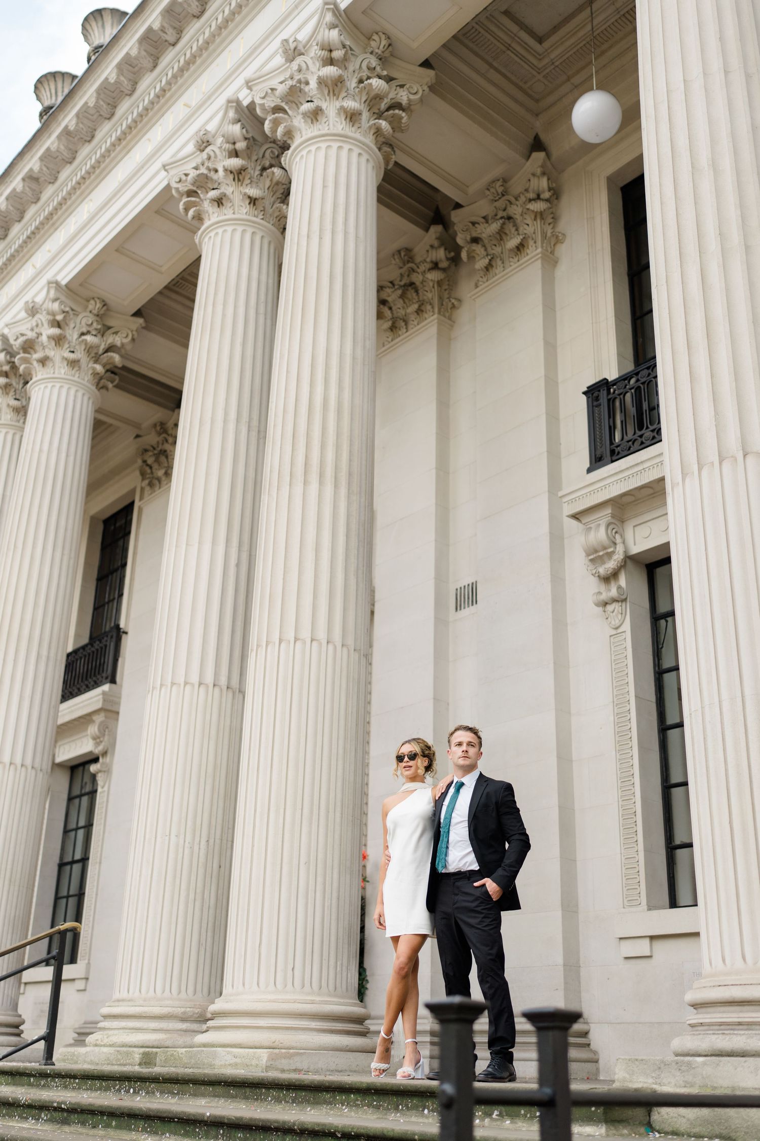 A couple poses between grand white columns of a neoclassical building.