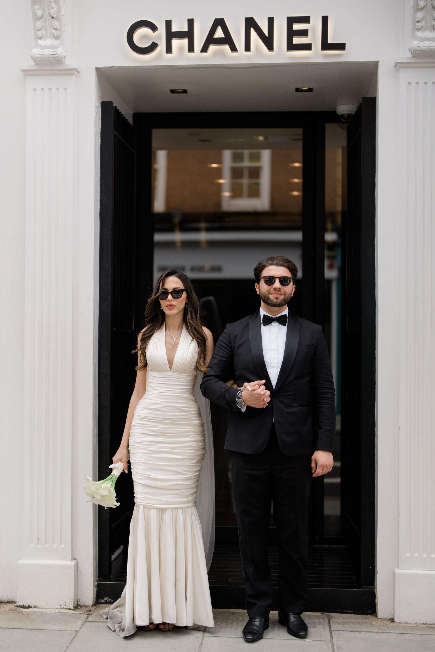 A couple poses outside a Chanel store entrance in formal wedding attire.