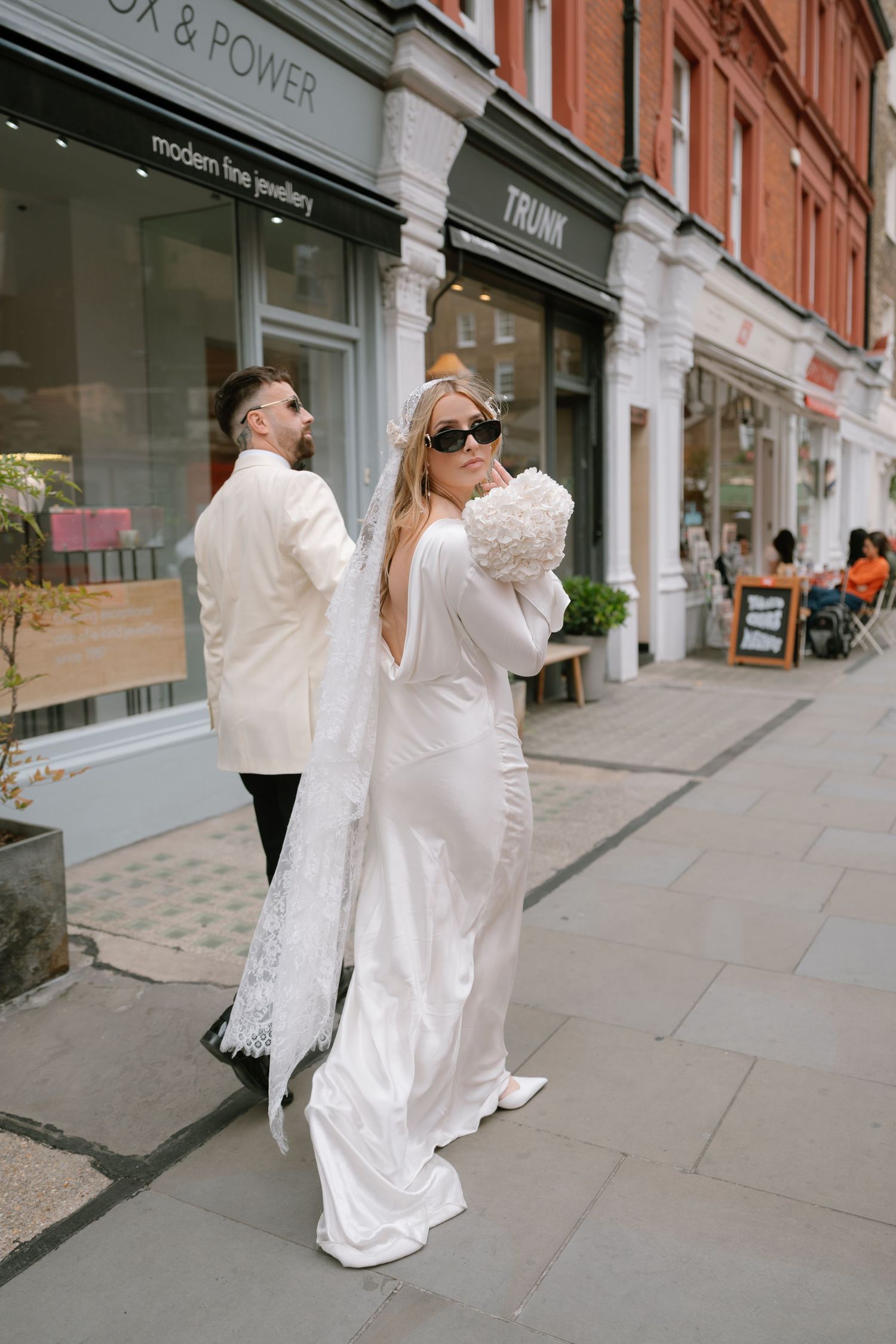 Couple in wedding attire walking past storefronts on urban sidewalk.