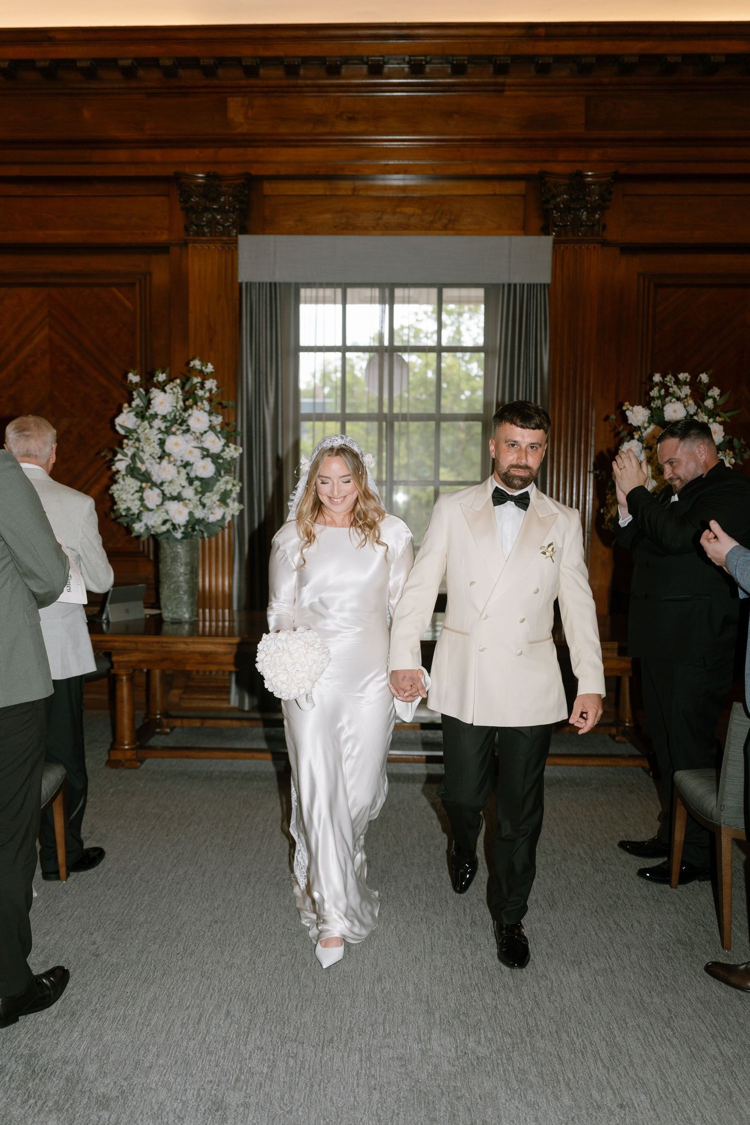 A newlywed couple walks down the aisle in a wood-paneled room decorated with white floral arrangements.
