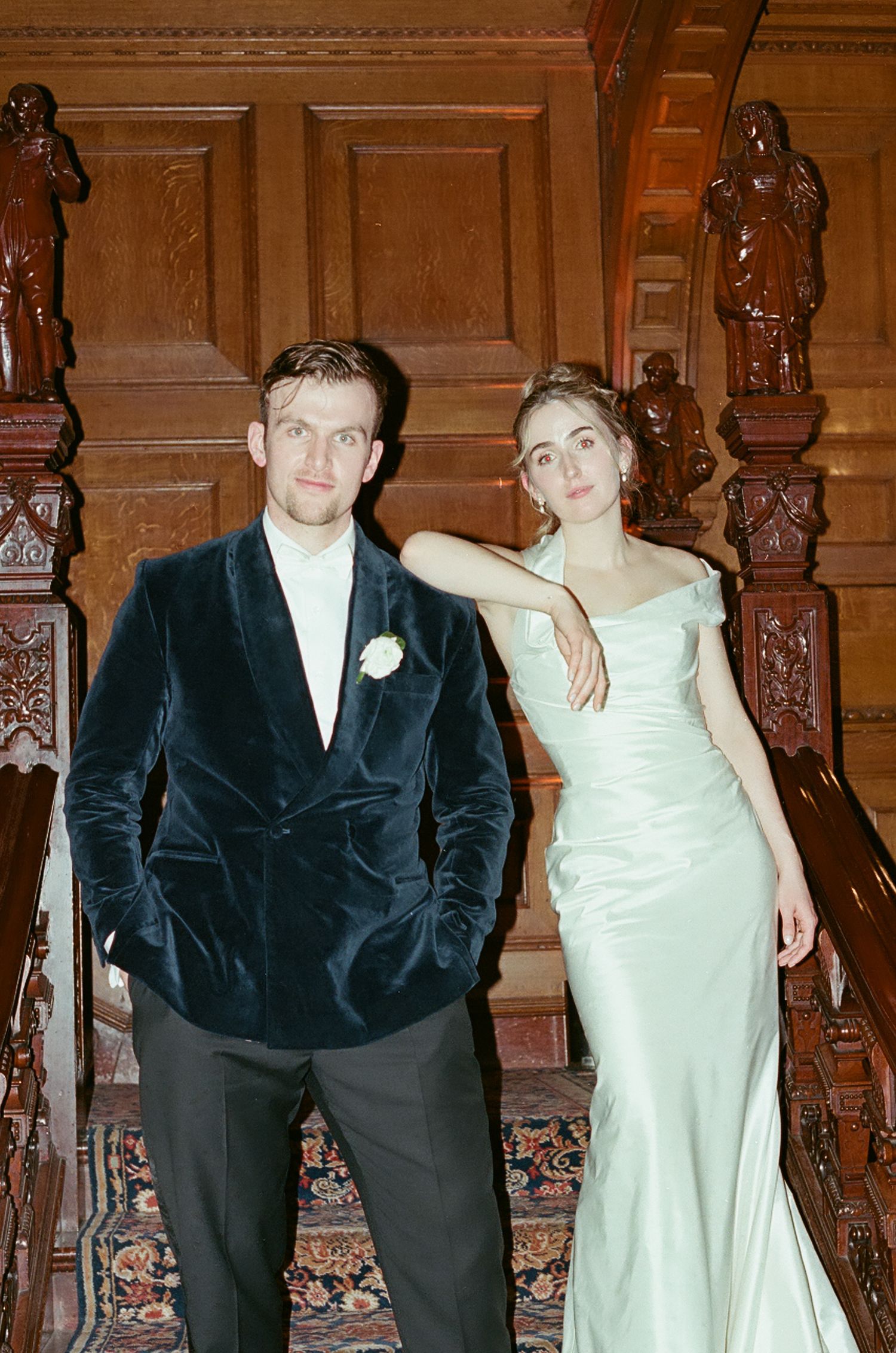 Group of people in formal attire pose against ornate wooden paneled wall with black suits and white dresses.