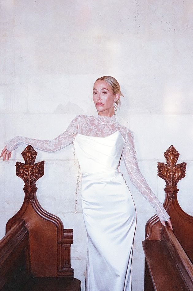 Bride in white long-sleeved gown poses gracefully against vintage wooden church pews in dramatic lighting.