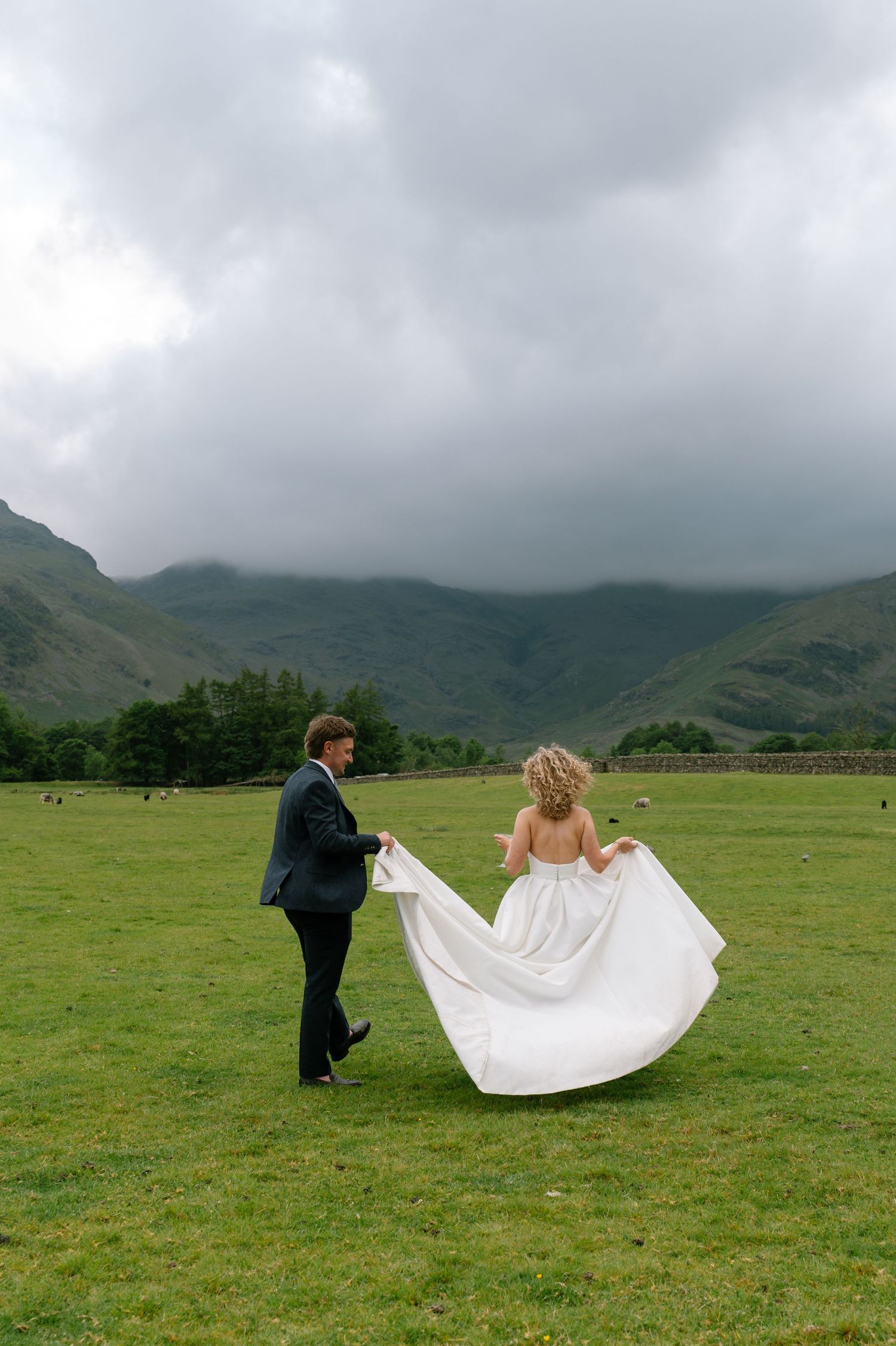 People arrange a wedding dress train in a green field with mountains in the background.