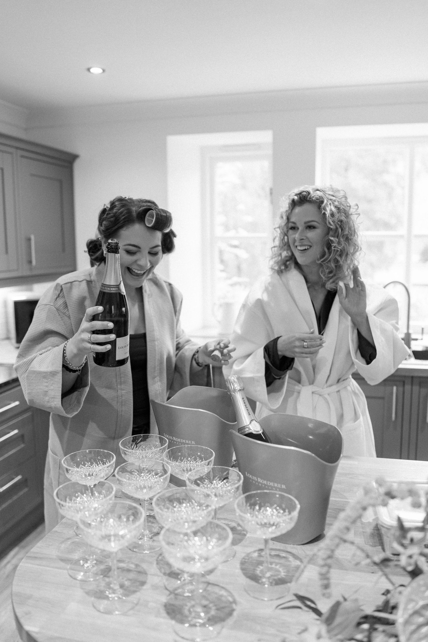 Two people in white robes prepare champagne glasses during wedding preparations in a bright kitchen.