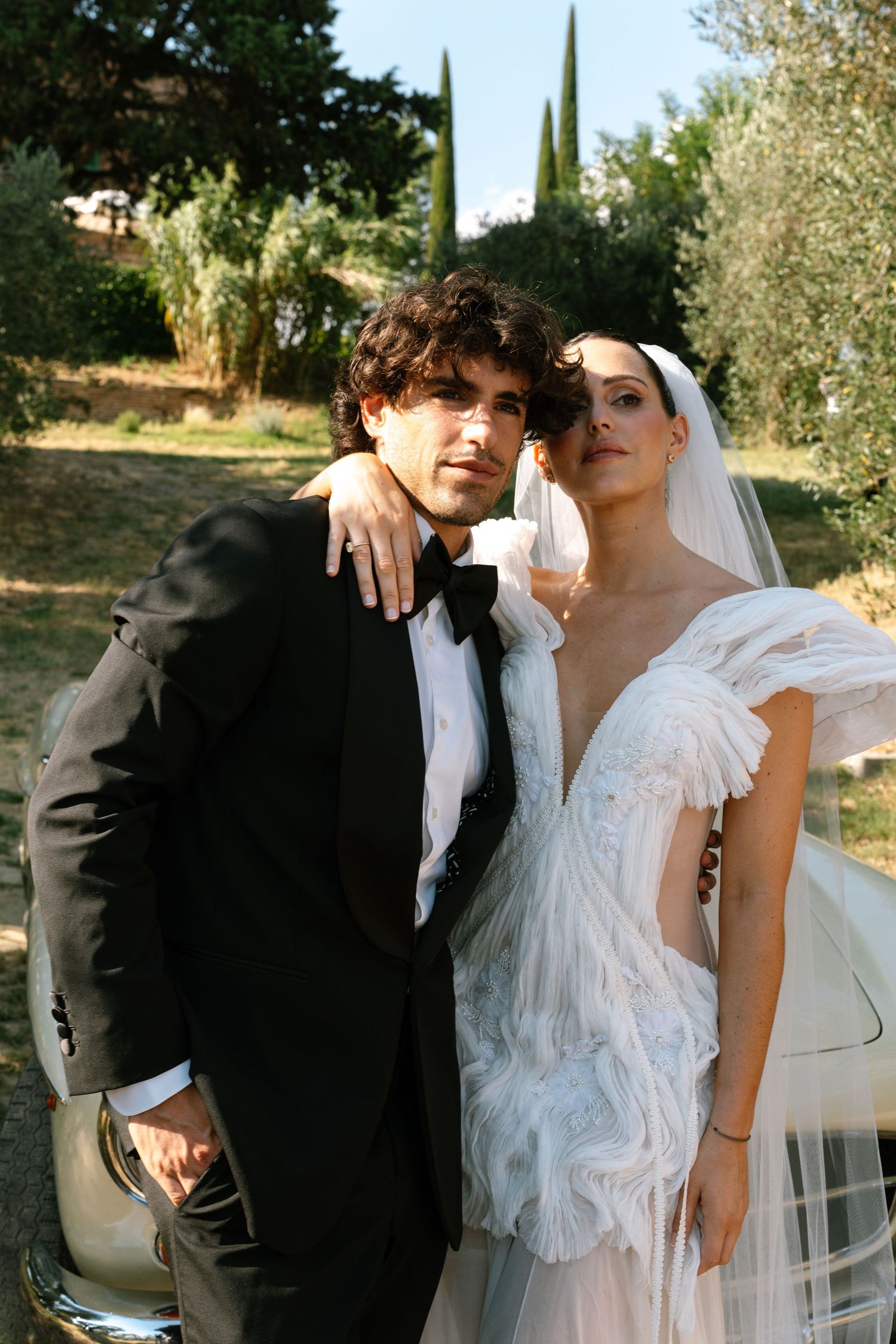 A newlywed couple poses together against a backdrop of olive trees and cypress.