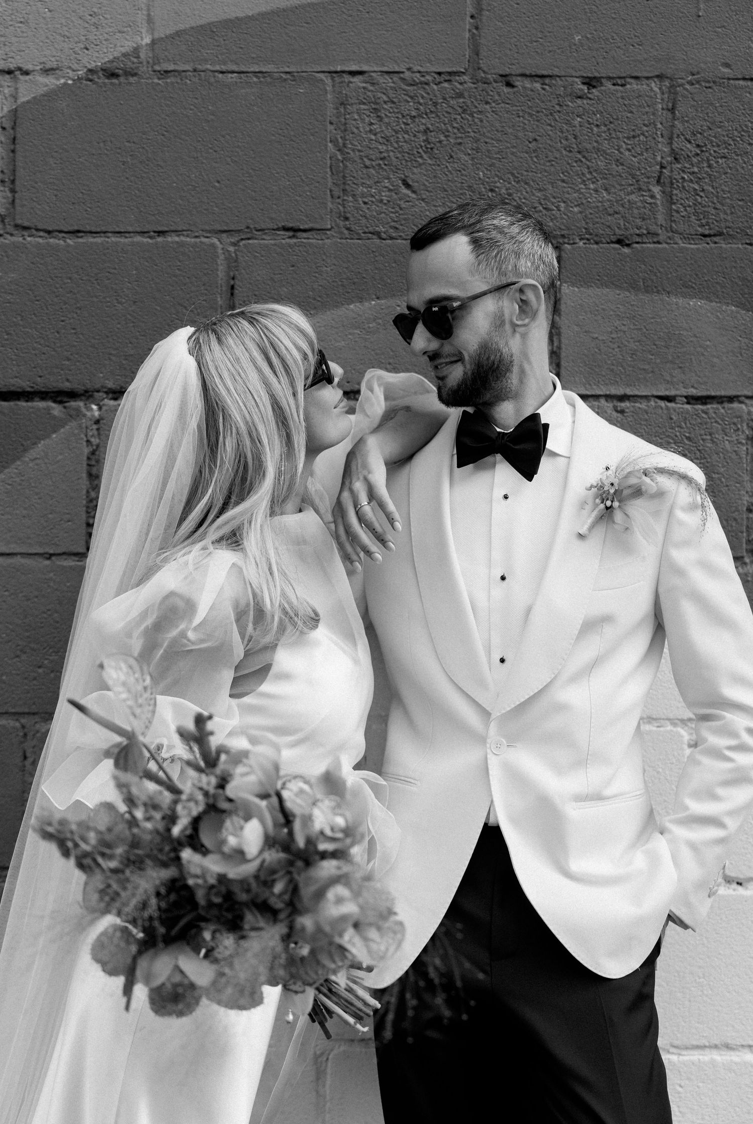 A couple in white formal attire poses against an industrial brick wall with floral bouquet.