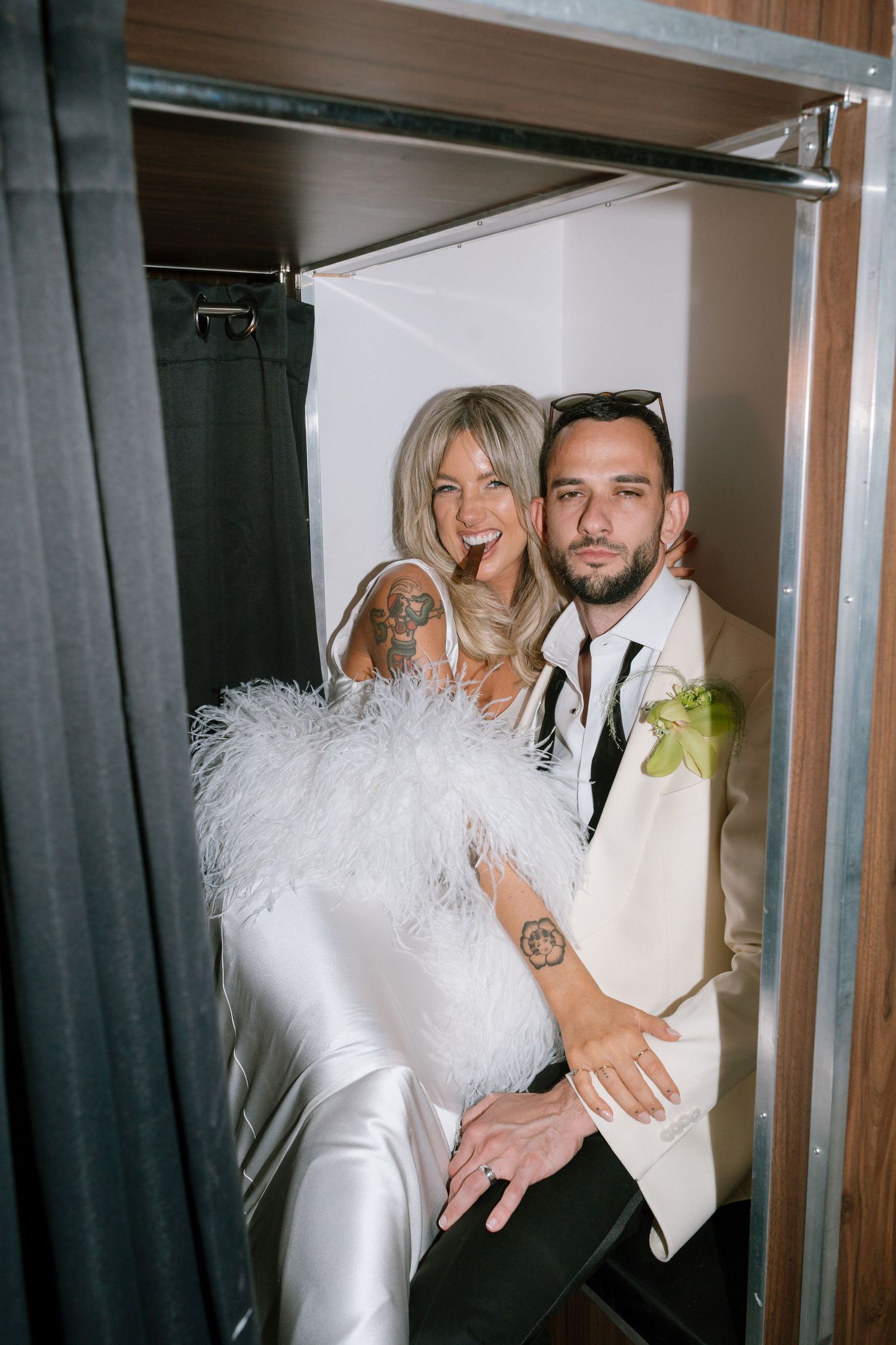 A couple poses in a white wedding outfit and suit against dark curtains in an intimate indoor setting.