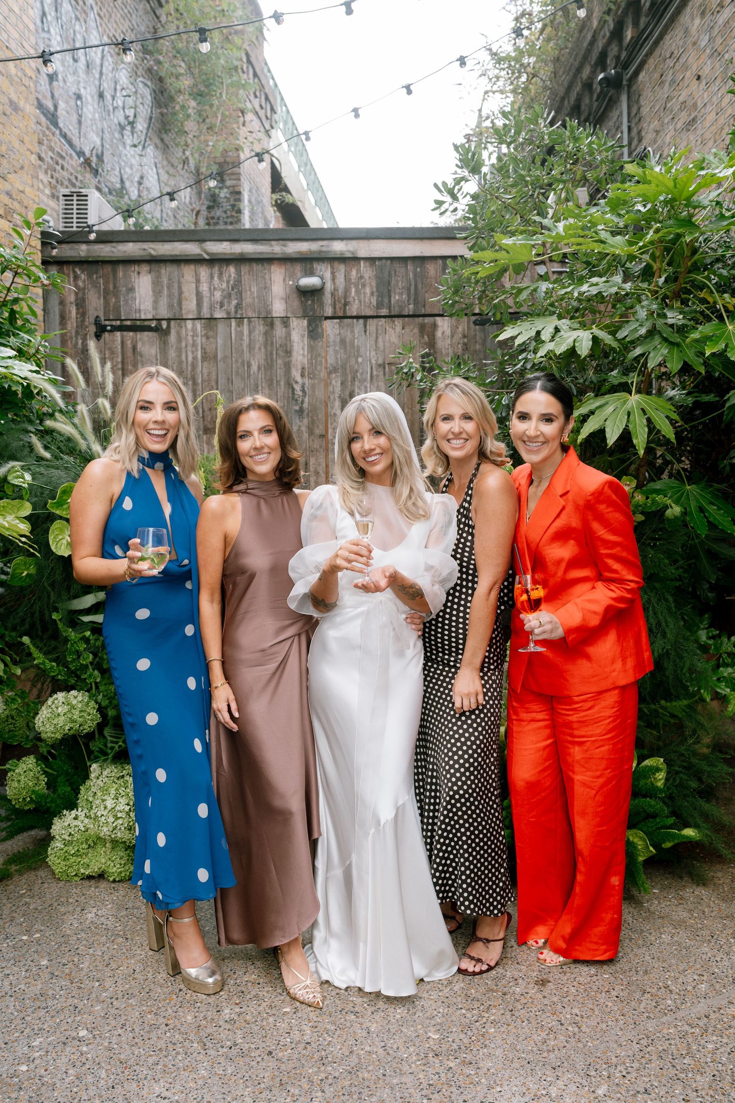 Wedding party in colorful dresses posing together against stone wall backdrop in garden setting.