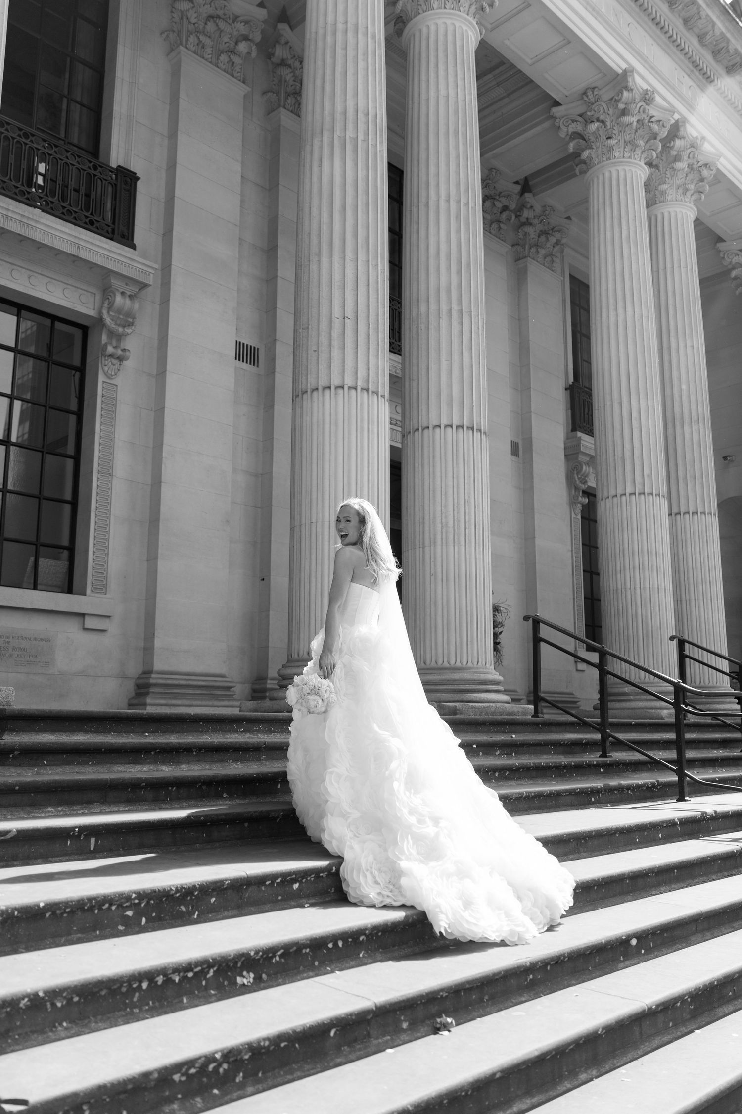 Bride in elegant white gown and veil poses on steps of classical building with tall columns, black and white photo.