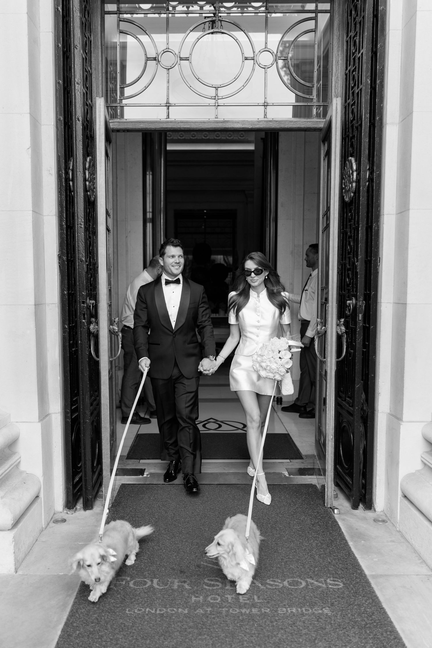 Elegant couple in formal attire walking two small dogs outside Four Seasons London at Tower Bridge in black and white.