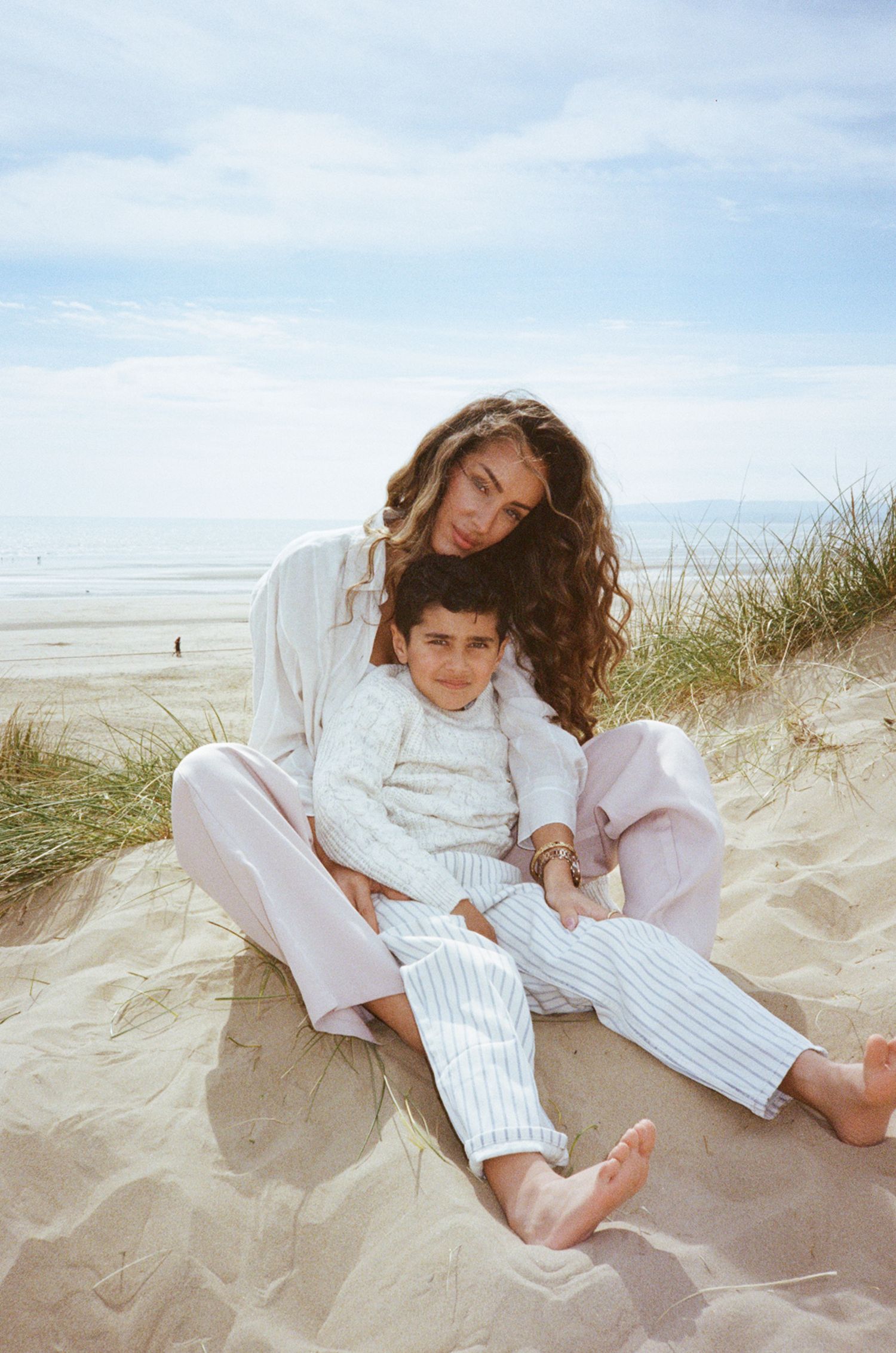 A woman and child sit together on sandy beach dunes, both wearing light pastel pajamas, with ocean waves visible behind them.