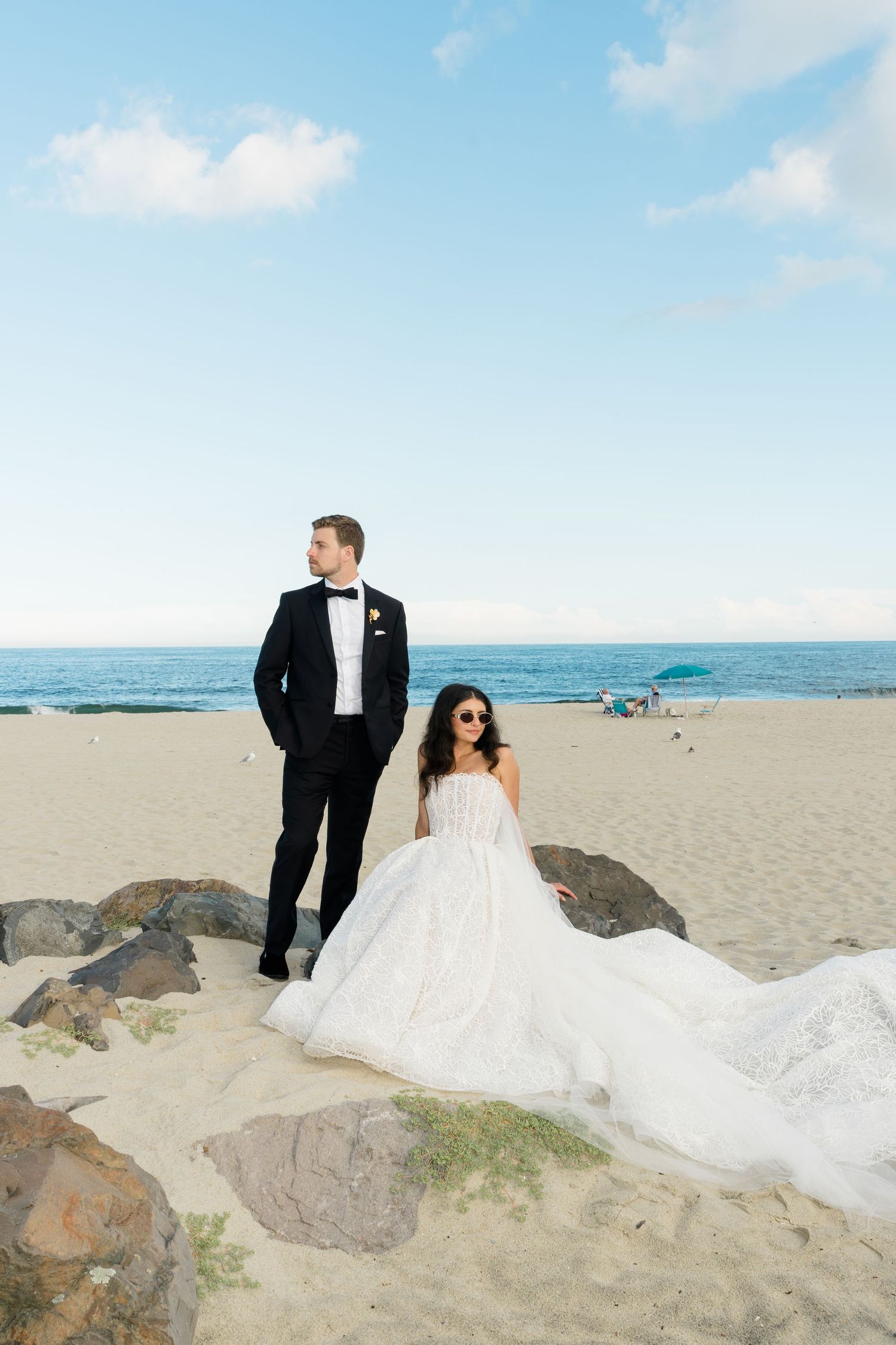 Bride in white ball gown and groom in black tuxedo pose on sandy beach with ocean backdrop.
