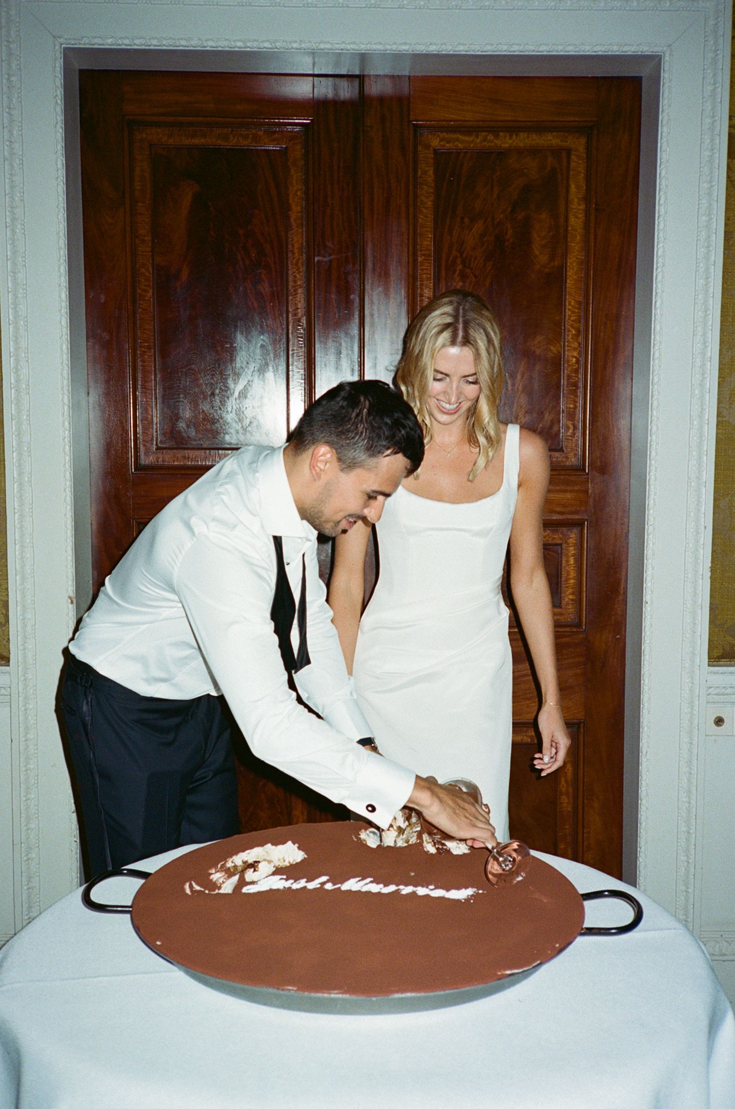 Newlyweds cutting a chocolate Just Married cake together, bride in white dress, groom in white jacket, wooden doors backdrop.