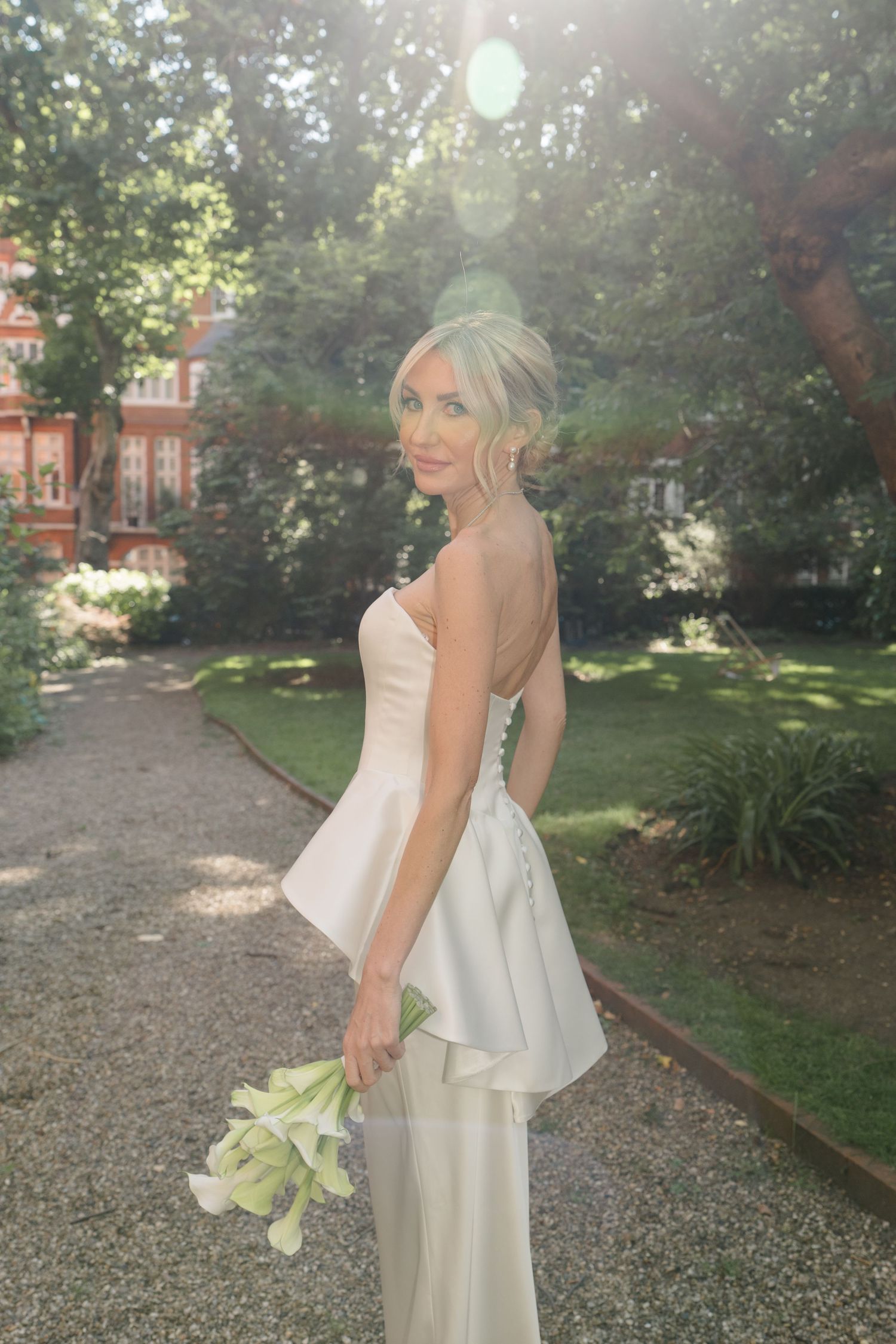 Bride in strapless white gown holding calla lilies, posing in sunlit garden with lush greenery and brick building.