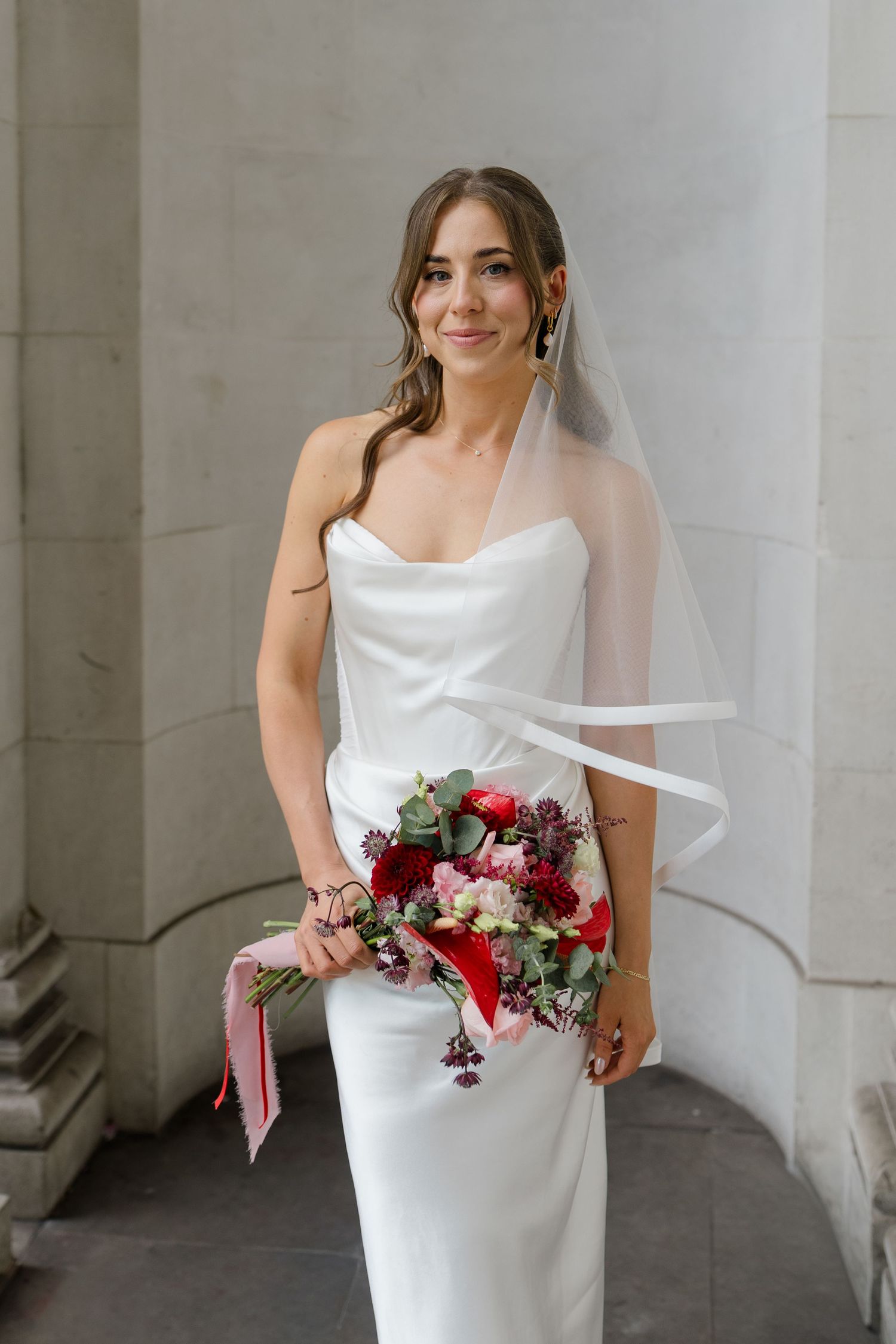 Bride in strapless satin gown holds red and pink bouquet with ribbon while posing between stone columns.